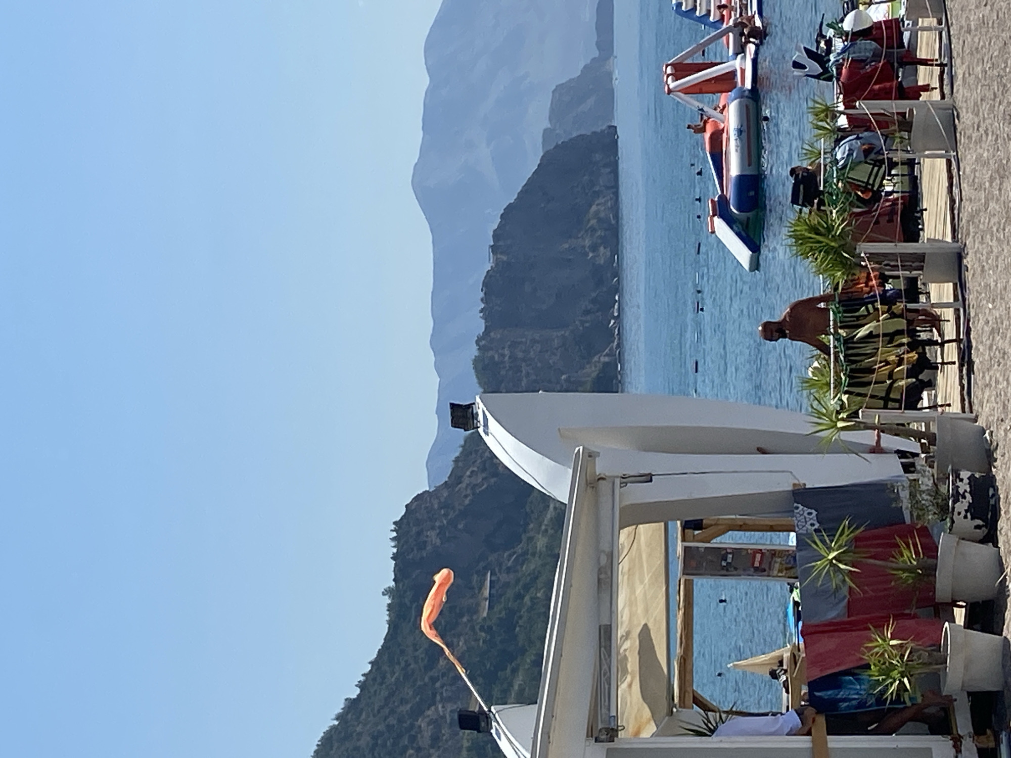 This image captures a vibrant scene at a beachside establishment near Prijevor, Montenegro. In the foreground, a white structure, possibly a bar or beach hut, is partially visible. Colorful towels and laundry are draped over a railing in front of the structure, with potted plants adding a touch of greenery.
Beyond the beach hut, the azure waters of the Adriatic Sea stretch out, dotted with people swimming and enjoying themselves. A large inflatable water park with slides and platforms is a prominent feature on the water.
In the background, majestic mountains rise from the coastline, their slopes covered in lush greenery. The sky is a clear, bright blue, indicating a sunny day. The overall impression is one of a lively summer day at a scenic coastal destination.