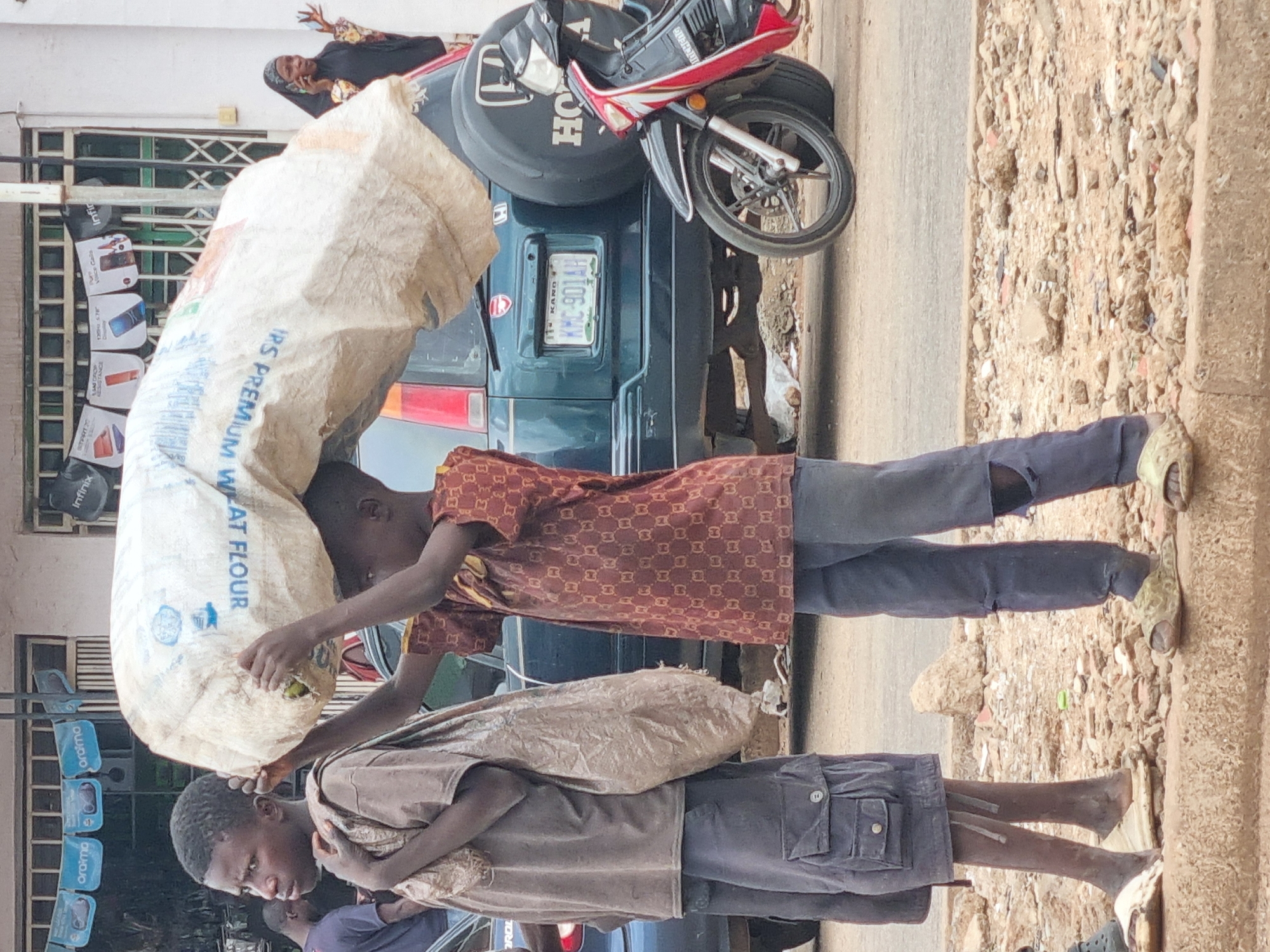 In Jos, Nigeria, two young boys are pictured on a roadside. The boy on the right, wearing a brown patterned shirt and torn blue trousers, carries a large, white sack of flour over his head. The boy on the left, in a grey t-shirt and blue shorts, walks beside him, carrying a smaller, lighter-colored bag slung over his shoulder. They are standing on a raised concrete curb overlooking a dirt and rubble-strewn area next to a paved road. Behind them, a dark green SUV with a spare tire mounted on the back is parked. A red and black motorcycle is parked beside the SUV. In the background, a woman in a black hijab and a patterned black and yellow garment is visible, holding a cell phone to her ear. A building with barred windows and various signage, including advertisements for cell phones, is situated behind the woman. The overall scene depicts a typical street environment in the city.