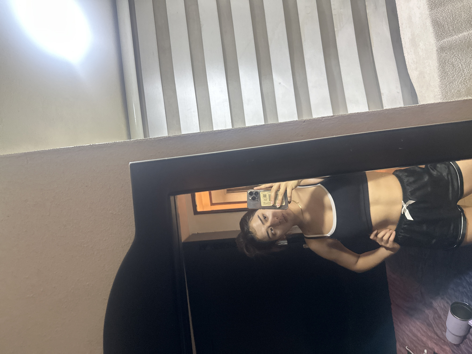 The image shows a woman in a black and white sports bra and black shorts taking a mirror selfie. The reflection shows a room with vertical blinds. A cup is visible in the lower right corner. The location is presumed to be indoors, possibly in Puebla, Mexico, based on the request context. The woman is holding a smartphone, likely taking the photograph.