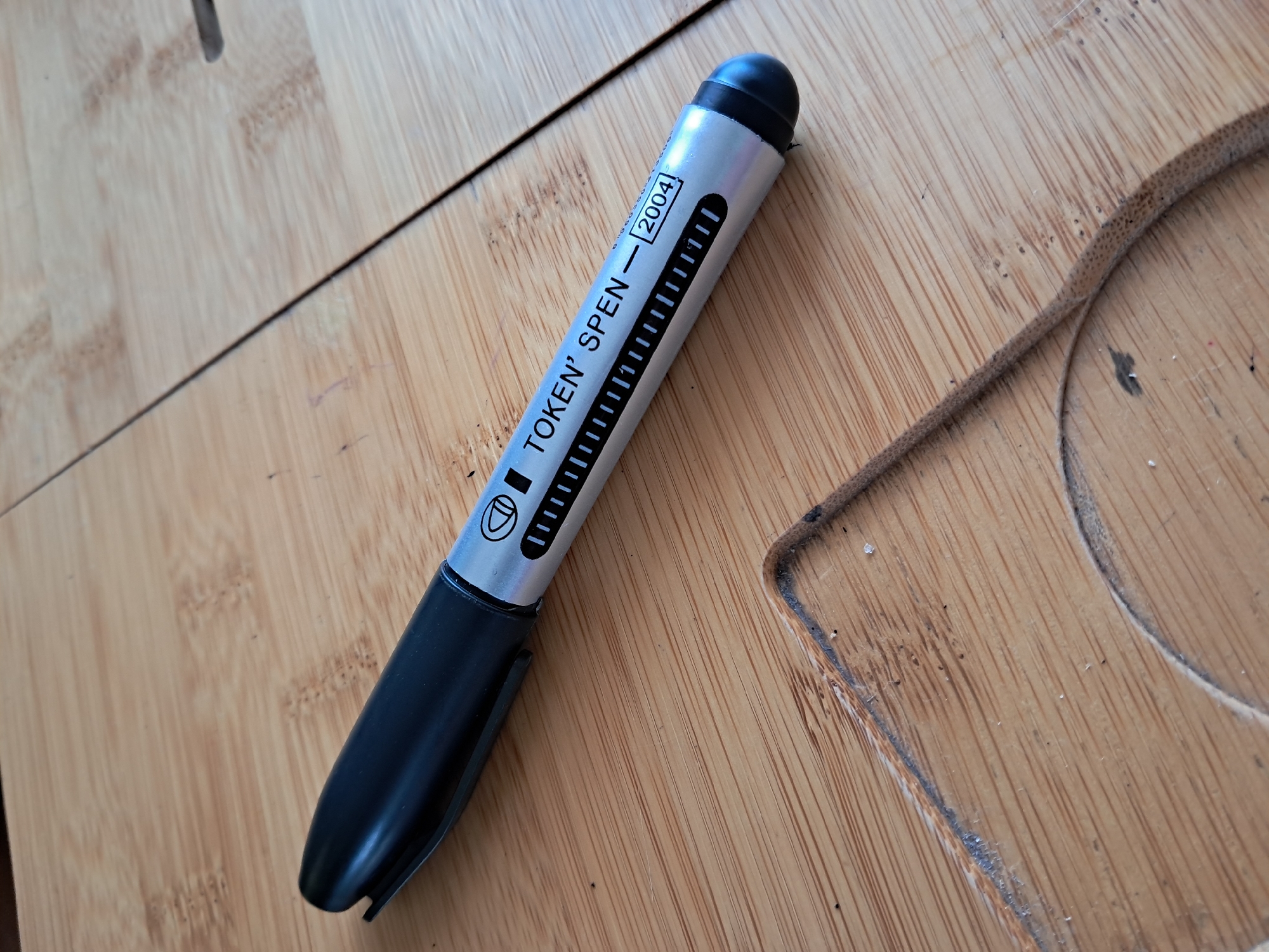 An eye-level shot captures a silver and black stylus or pen, labeled "TOKEN' SPEN - 2004", resting diagonally on a light-colored wooden surface. The stylus has a black rubber tip at one end and a black cap with a clip at the other, suggesting it can be attached to a pocket. Below the text "TOKEN' SPEN", a series of small, vertical hash marks are printed on the silver barrel. To the left of "TOKEN'", a small circular icon with a downward-pointing arrow or stylized "D" is visible.
The wooden surface appears to be made of bamboo or a similar light wood, with distinct parallel grain patterns. There are faint lines and minor imperfections on the wood, indicating wear. To the right of the stylus, a curved, irregular cut-out or groove is visible in the wooden surface, possibly part of a larger design or functional element like a holder. A thin, dark brown, textured strip, possibly made of leather or rubber, is partially visible along the lower right edge of the frame, lying on the wood. The lighting is bright and even, suggesting an indoor setting during the daytime. The scene is set in Kyiv, Ukraine.