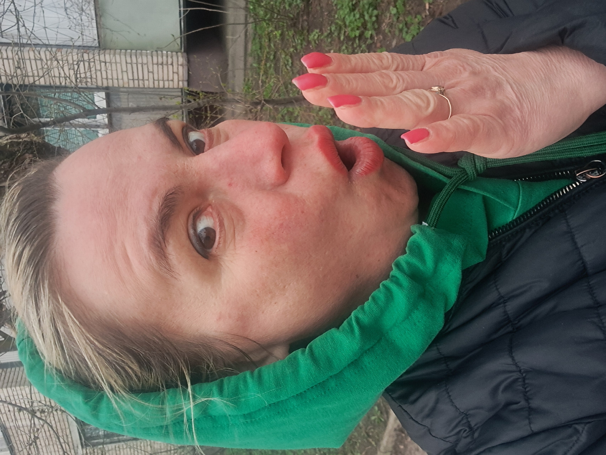 A woman with fair skin, blonde hair, and pink nail polish poses for a close-up shot outdoors. She wears a green hooded sweatshirt under a black quilted jacket. Her mouth is puckered and her eyes are wide. She holds her right hand up near her face, displaying a gold ring on her ring finger. The background shows a brick building and greenery, suggesting an urban or suburban environment. The location is Elektrostal, Russia.