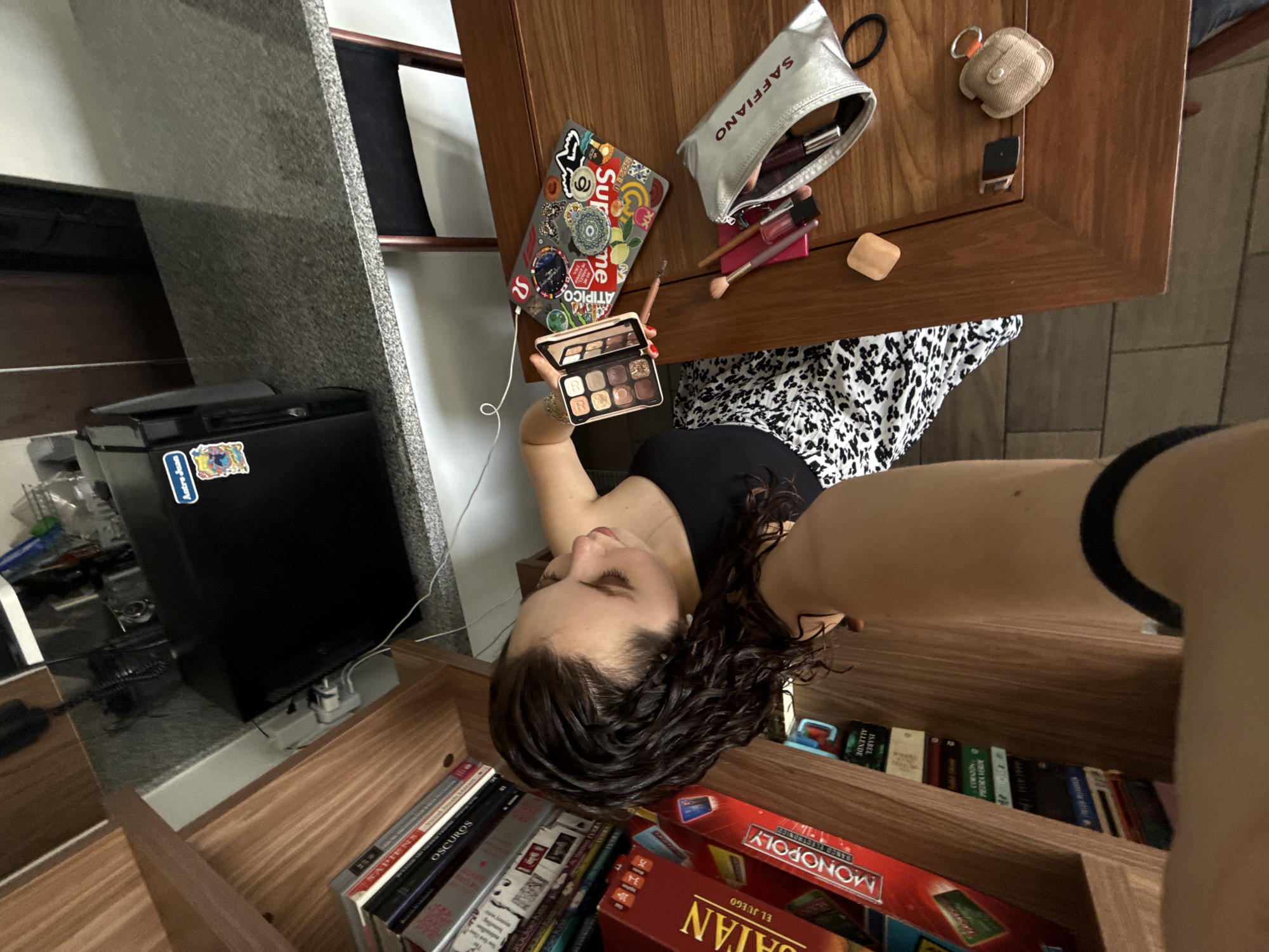 This high-angle, rotated selfie captures a casual indoor scene, likely in a home in Mexico City, Mexico. The primary subject is a person, appearing female, with dark, curly hair, wearing a black top and a black and white patterned skirt. Her face is turned downwards, partially visible, as she holds an open eyeshadow palette with multiple shimmer shades in her right hand. Her left arm extends into the frame, suggesting she is holding the camera.
The immediate foreground features a wooden table laden with personal items. A silver cosmetic pouch, labeled "SAFFIANO," is open, revealing various makeup items like lip glosses and brushes. Beside it rests a gray laptop or tablet decorated with numerous colorful stickers, including one that reads "Supreme" and another "ATIPICO," with a white charging cable attached. A small, light brown fabric pouch, possibly for earbuds, and a small black object are also on the table.
In the background, to the left, a black mini-fridge with a sticker partly visible as "Antro Juan" sits next to a gray, speckled countertop or wall. Below the table and to the right, a wooden shelf unit is filled with books and board games. Visible book titles include "SAPIENS," "OSCUROS," and "ISABEL ALLENDE." Board game boxes are also present, with "MONOPOLY" clearly identifiable and another red box showing elements consistent with "CATAN" (partially visible as "UATAN" in OCR, with "EL JUEGO" below). The floor, visible in areas, appears to be tiled.
The scene depicts a quiet, personal moment, possibly of the individual preparing for makeup application or simply examining the palette. The lighting suggests daytime.