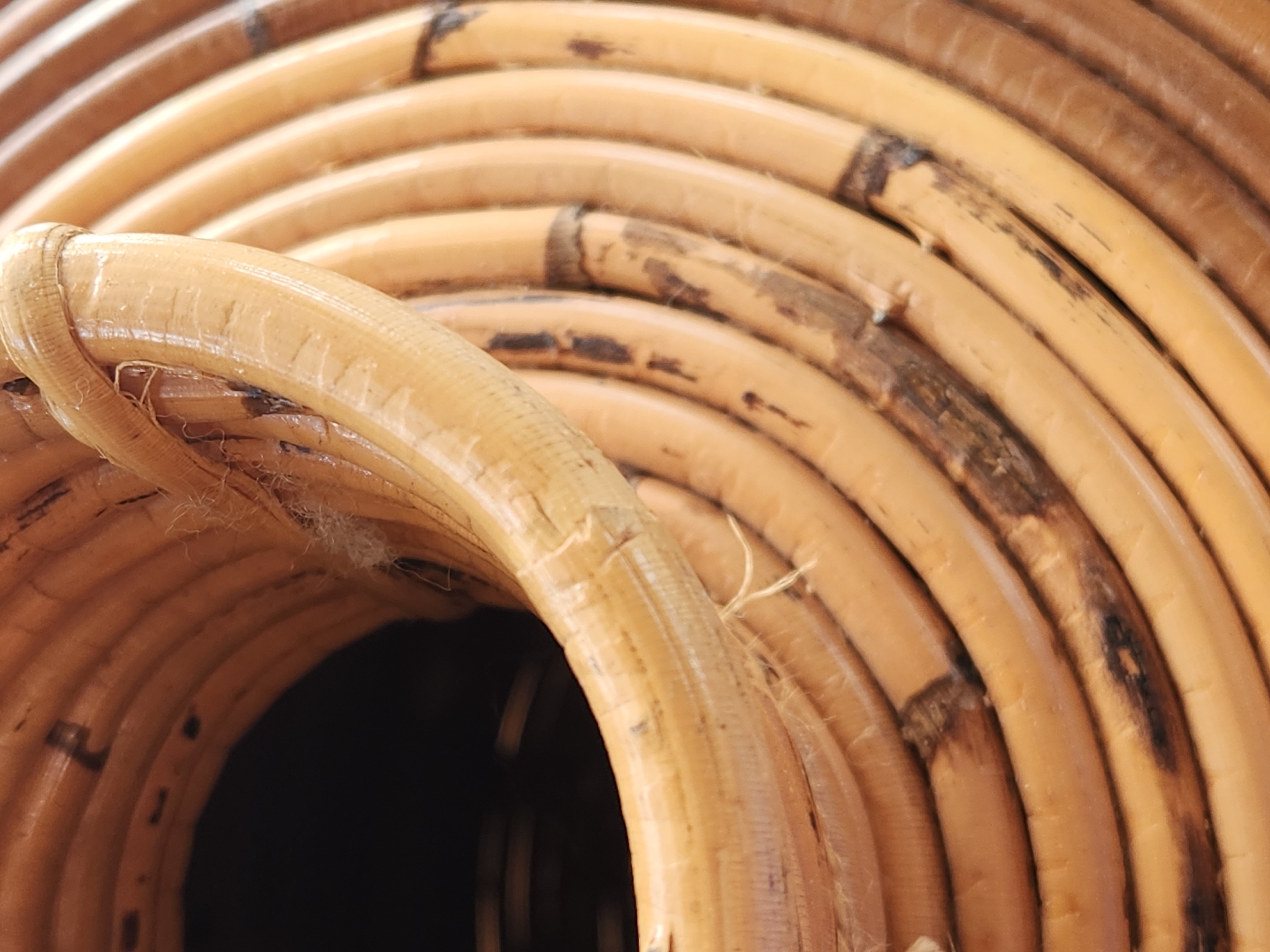 This is a close-up, abstract photograph of tightly coiled wicker material, likely from a basket or furniture. The dominant subject is the texture and pattern of the wicker, which is a natural, light brown color with darker, charred-looking imperfections and variations in tone. The wicker strands are rounded and smooth to the touch, with some appearing slightly glossy. The coils create a repetitive, spiral pattern, drawing the viewer's eye inward towards a dark, shadowed opening at the center. Fine, wispy fibers are visible, particularly on the outermost coil on the left, adding to the organic and handmade feel of the material. There is no visible text, people, or discernible activity or event taking place. The lighting appears to be soft and diffused, suggesting an indoor setting or an overcast day, though no specific location cues are present beyond the given context of Maitland, USA. The overall impression is one of natural materials and craftsmanship, with a focus on texture and form.