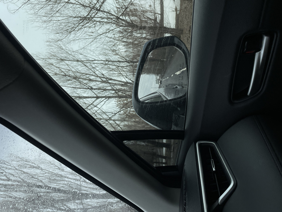 The image presents a view from the interior of a vehicle, likely in or near Solak, Armenia. The perspective is directed towards the front passenger side, encompassing the vehicle's dark dashboard with a chrome-trimmed air vent, a dark door panel with a chrome handle, and the A-pillar. The exterior window is visibly speckled with raindrops.
Through the window and reflected in the side-view mirror, a winter or early spring landscape is visible under an overcast, light grey sky. Numerous bare deciduous trees, stripped of leaves, line the roadside. The side mirror clearly reflects a two-lane paved road with white lane markings, where a grey sedan is observed directly behind the camera's vehicle, and a truck is visible further down the road in the distance. The mirror's surface displays the warning "OBJECTS IN MIRROR ARE CLOSER THAN THEY APPEAR." To the right of the road, beyond the trees, the ground shows patches of snow or ice, bordering a body of water. No individuals are visible in the scene.