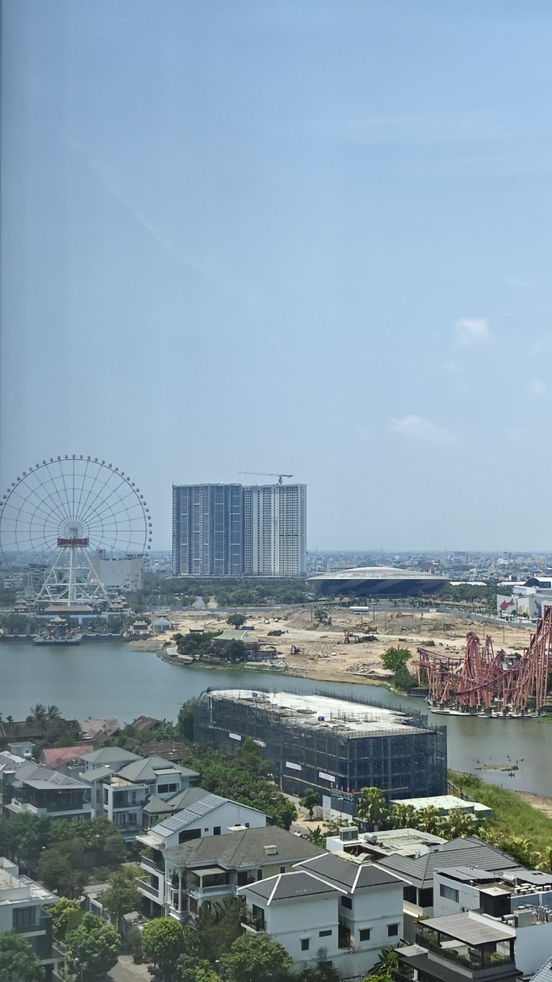 The image shows an elevated daytime view of a cityscape. In the midground, there is a body of water with a ferris wheel on the left, a tall skyscraper in the center, and a large stadium slightly to the right. There is construction work taking place in the area between the water and the structures. In the foreground, there is a residential area with several houses surrounded by trees. The image is likely taken in Đà Nẵng, Vietnam, based on the urban landscape, architectural style, and presence of a ferris wheel, a stadium and a highrise buildings. There are no people visible in the image. The sky is light blue with a few clouds.
