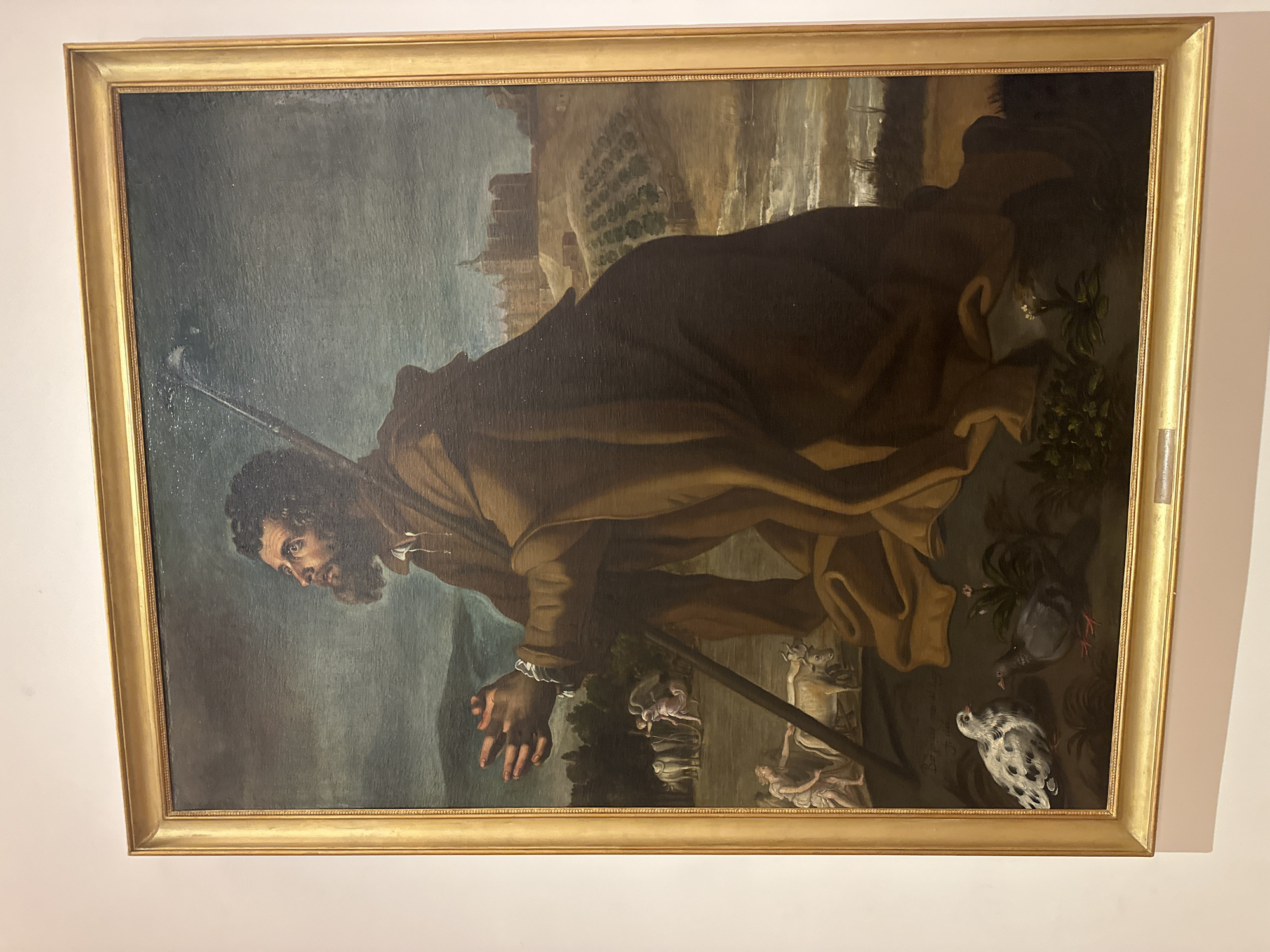 A painting depicts a man with a beard and dark hair kneeling with his hands clasped in prayer, looking upwards. He wears a brown robe and holds a staff. In the background, a town with a walled structure and a church is visible on a hill, with agricultural fields in the middle ground. A river flows in the foreground, and there are two birds, a speckled dove and a grey pigeon, in the lower left. There are also figures and animals, including cows and an angelic figure, in the middle ground. This painting is located at the Prado Museum in Madrid, Spain.