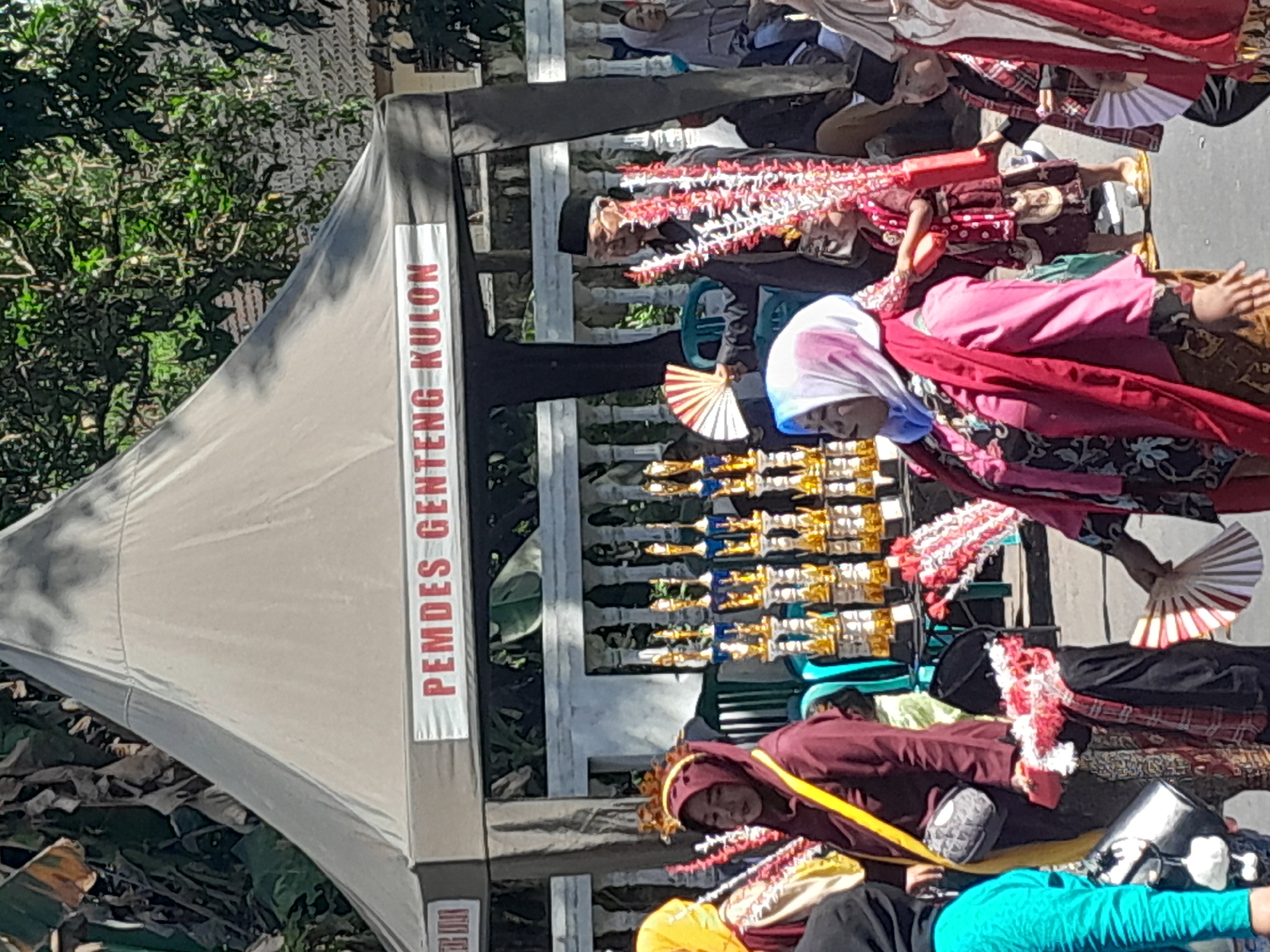 The image captures an outdoor scene at an event, likely a festival or competition, in Genteng Kulon, Indonesia. A large, beige tent with the words "PEMDES GENTENG KULON" printed in red on a white banner dominates the background, indicating the location. In front of the tent, several rows of trophies, predominantly gold and blue, are displayed on black stands.
Several people are visible, dressed in traditional attire and participating in the event. A woman in the foreground on the left wears a maroon headscarf and a yellow sash. To her right, a woman in a pink headscarf and a bright pink outer garment is holding a red fan. Behind her, a young boy in traditional clothing is also holding a fan. Further to the right, a man in a black cap and traditional clothing is holding a fan and a decorative item with red and white elements. Other individuals, including children and adults, are also present, some seated and others standing, contributing to the lively atmosphere. The environment suggests a sunny day, with shadows cast by trees visible in the background.