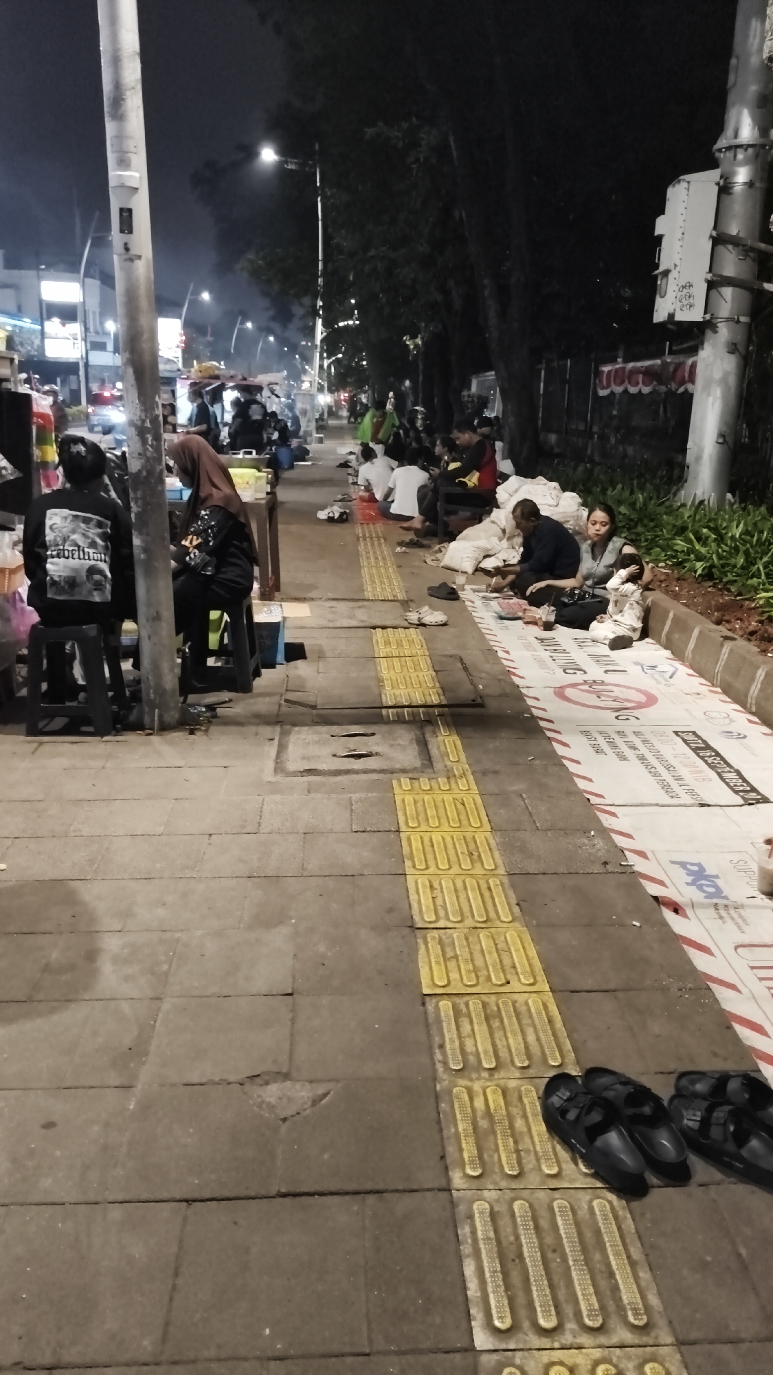 The image depicts a street scene at night in South Jakarta, Indonesia. A sidewalk is lined with people, some sitting on stools and others on the ground. Many appear to be eating or socializing. Food stalls are visible in the background, with smoke emanating from them, suggesting cooking is in progress. There are trees lining the sidewalk, and streetlights illuminate the area. The ground is paved with concrete tiles, and a tactile paving system is present. Several pairs of sandals are placed on the sidewalk. A large sheet with text and graphics is spread out on the ground in the foreground.