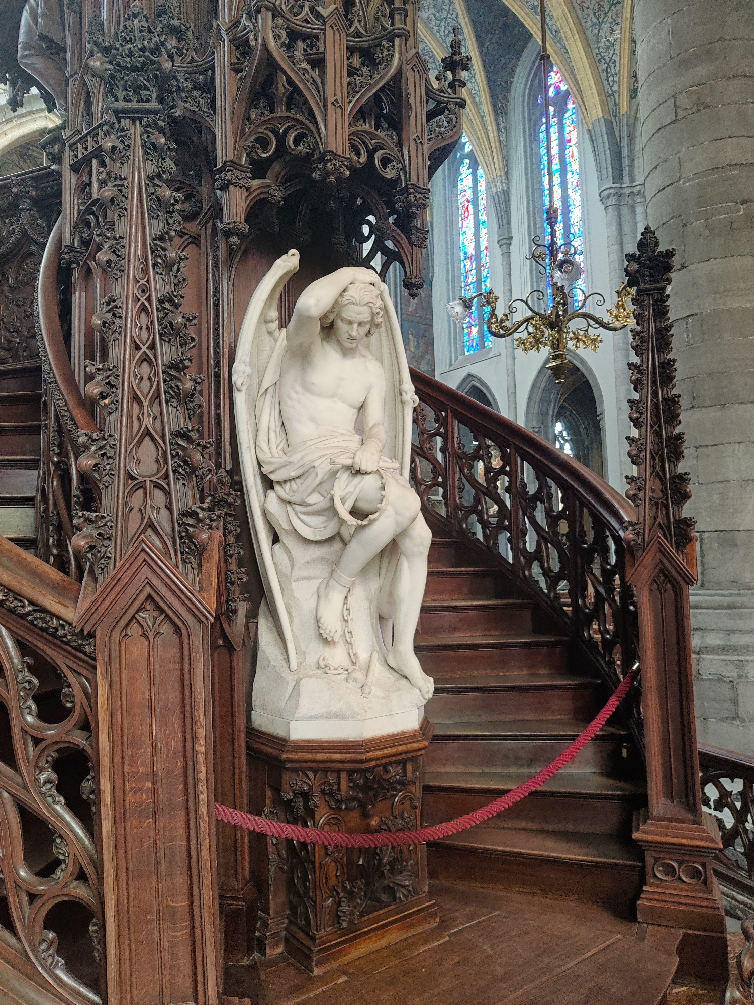 The image shows a white marble statue of a winged figure, possibly an angel, seated on a pedestal on a wooden spiral staircase inside a church in Liège, Belgium. The figure is looking down, with one hand on its head, and is holding a circular object, possibly a halo or a crown, in its lap. The staircase is ornately carved from dark wood, with intricate gothic-style detailing. Behind the staircase, stained-glass windows with blue and red panes are visible, along with a chandelier hanging from the ceiling. The floor is also made of wood. The overall scene suggests a place of worship or a historical building.