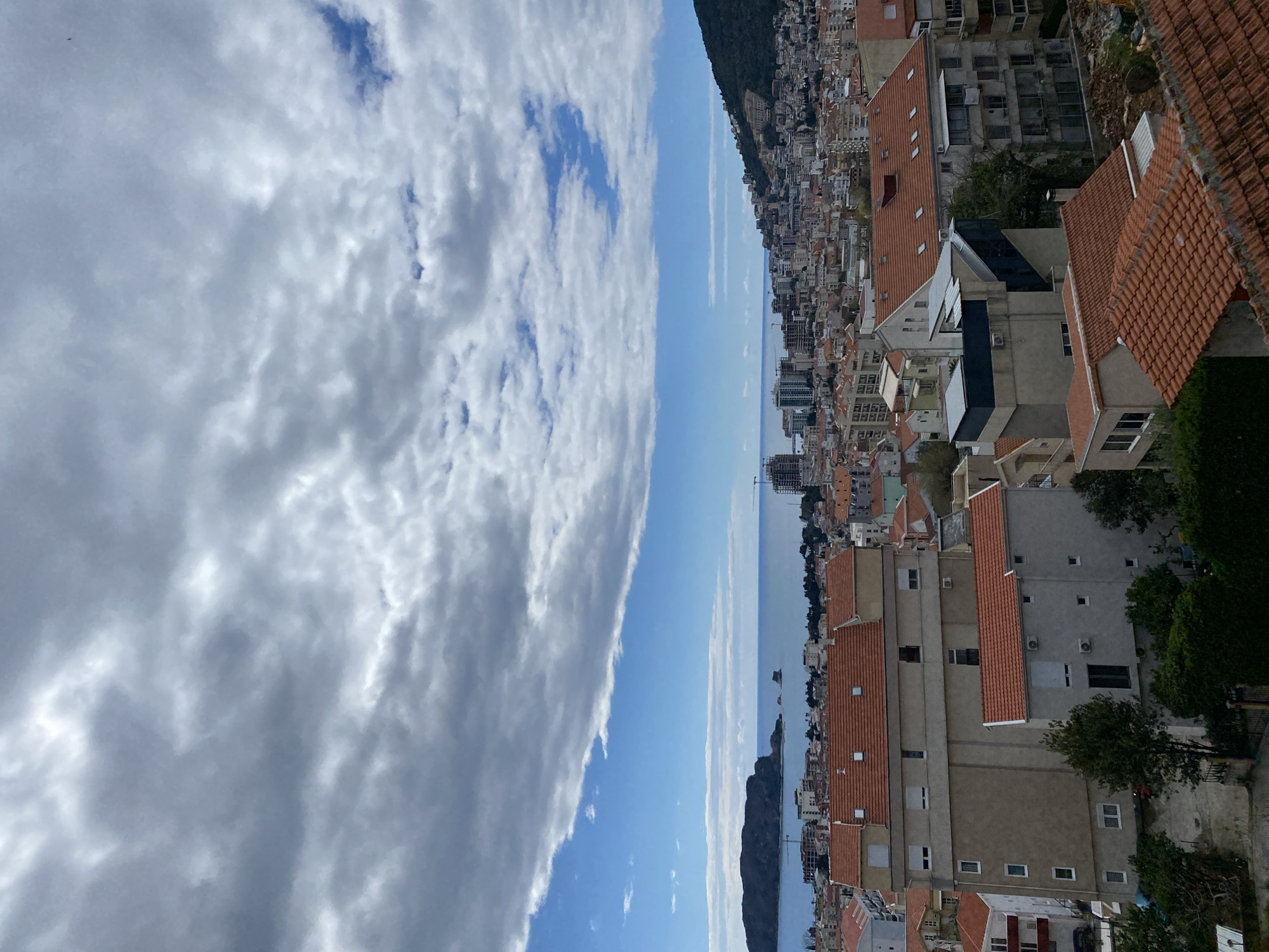 The image presents an elevated, slightly tilted view of a section of Budva, Montenegro. The right side of the frame is densely packed with buildings, many featuring red-tiled roofs, common to the region's architecture. The built environment extends towards the sea. Between the town and the sky is the body of water. The left side of the frame shows a dramatic sky, dominated by dense, grey, and white clouds. A sliver of blue sky breaks through the clouds.