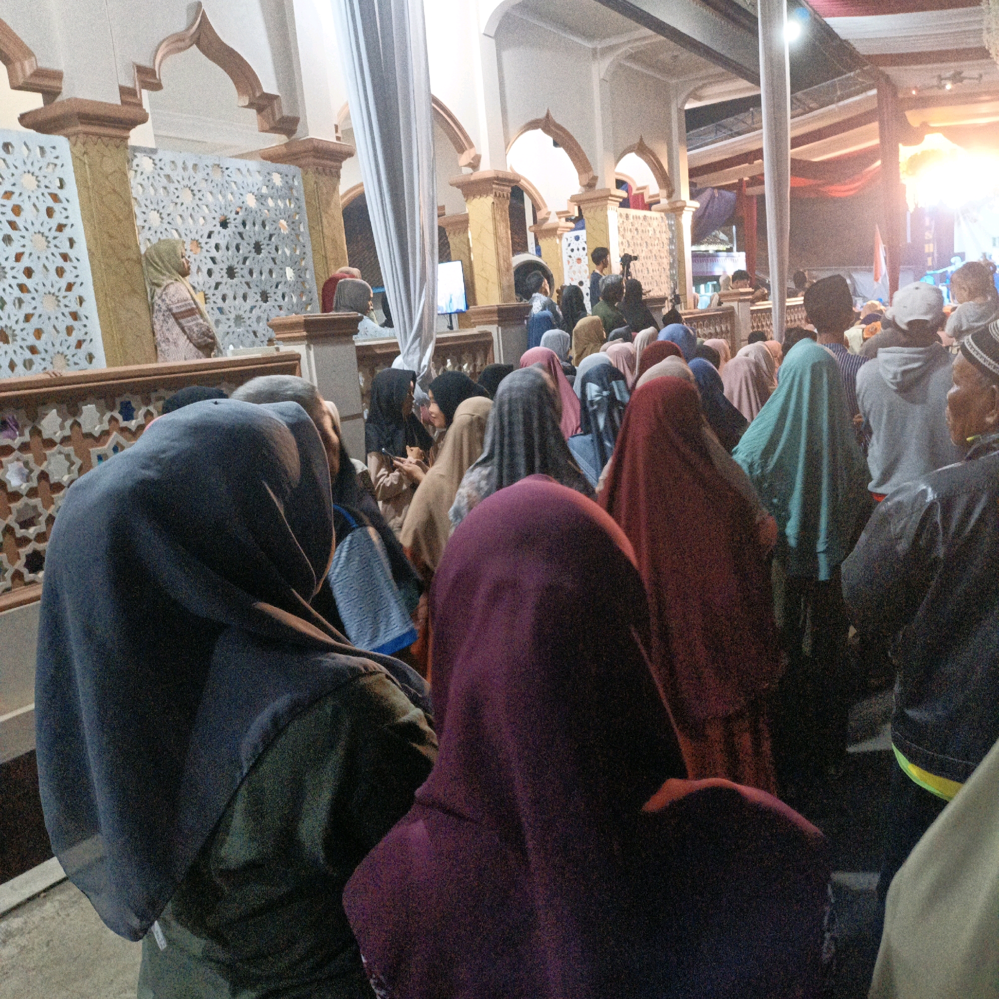 In the city of Kalitorong, Indonesia, a gathering of people, predominantly women wearing headscarves, are observed inside a building that appears to be a place of worship or a community hall. The architecture features arched doorways and ornate latticework. In the foreground, a woman in a dark headscarf faces away from the camera, her shoulders hunched. To her right, another individual's shoulder and headscarf are visible. Behind them, a crowd of people, many in colorful headscarves, are gathered. Some individuals are looking at a screen or stage area where a bright light suggests a performance or presentation is taking place. The overall scene depicts a communal event or religious observance.
