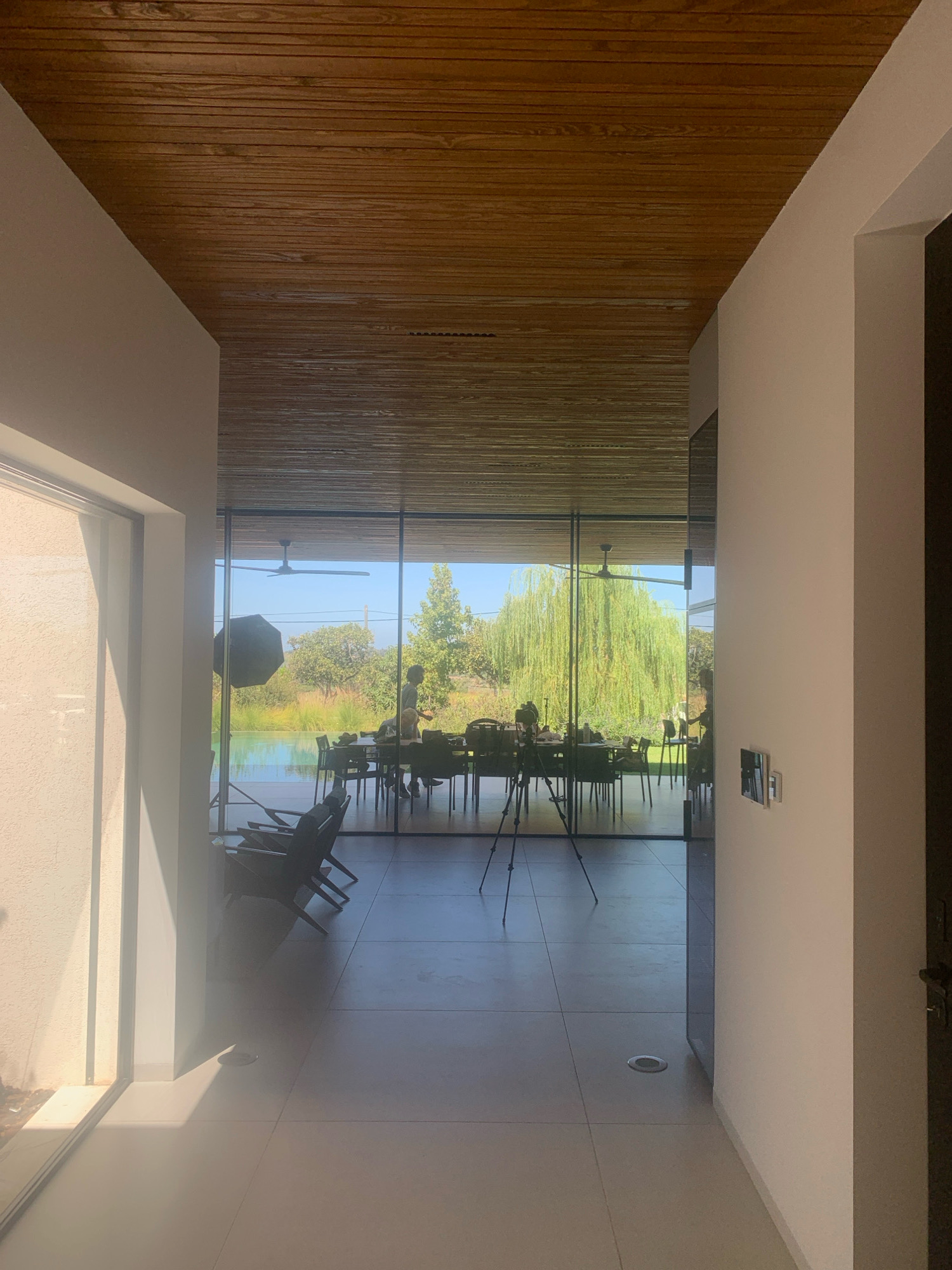 The image depicts a modern interior space with a large glass wall overlooking an outdoor environment. The ceiling is constructed from wooden slats, and the walls are painted white. The floor is tiled in a light color.
In the foreground, to the left, a large window lets in bright sunlight, casting strong shadows on the wall and floor. To the right, a white wall has a small silver framed object mounted on it. A dark doorway is visible on the far right.
Through the glass wall, a dining table with chairs is set up, suggesting an interior dining area or an event setup. Two people are visible near the table, one appearing to be a woman with blonde hair, and another individual. A large black umbrella-like object is positioned to the left of the dining area, possibly a photographic light modifier. A tripod with a camera is set up in front of the dining table.
Beyond the dining area, a swimming pool is visible, and the water reflects the sky and surrounding greenery. The landscape outside features trees, including a prominent weeping willow tree, and lush vegetation under a clear blue sky. The overall impression is a contemporary home or venue with a strong connection to its natural surroundings. The location is identified as Kfar Monash, Israel.