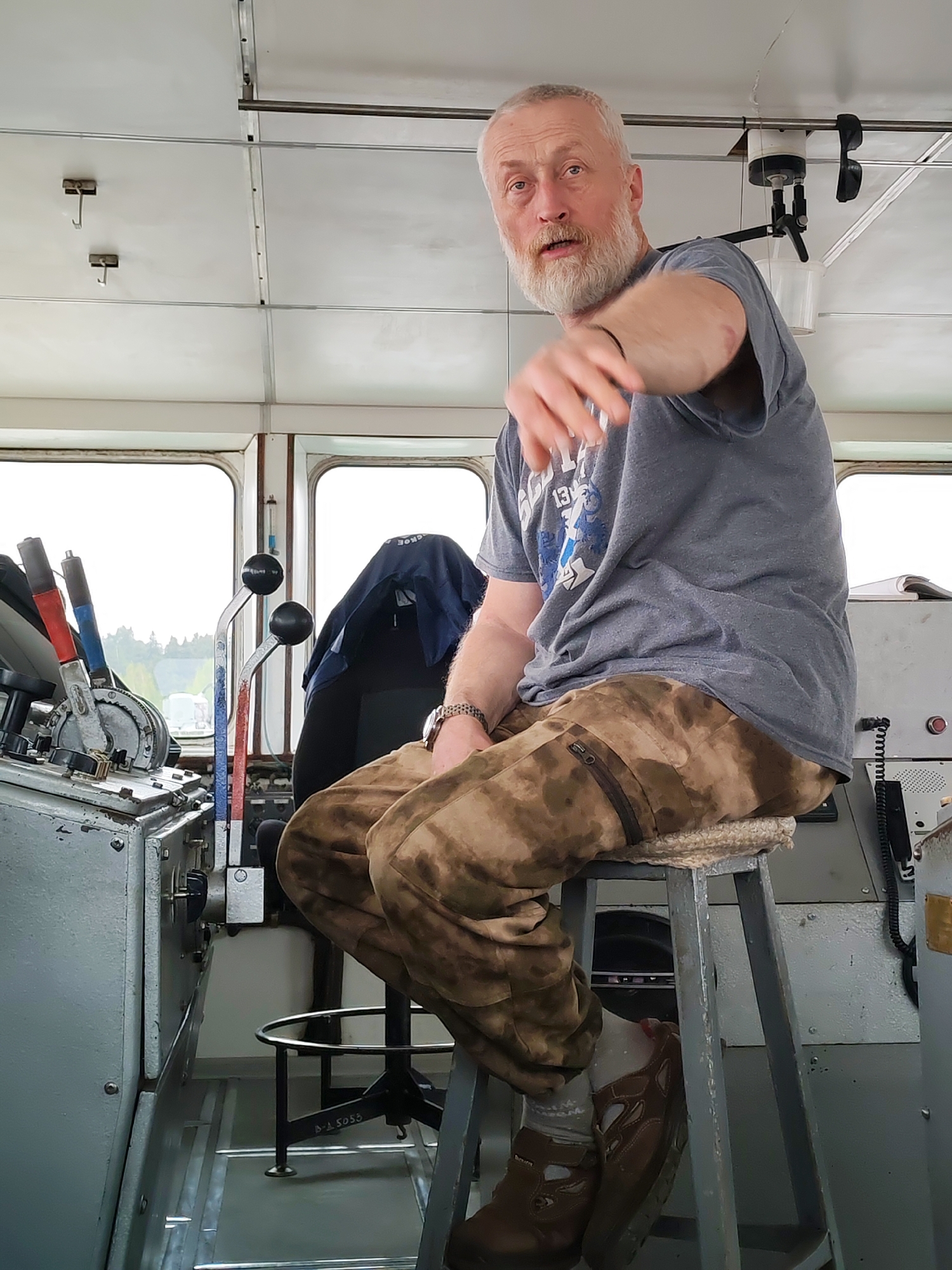 The image shows a man with a white beard sitting on a stool in what appears to be the control room of a ship. He is wearing a gray t-shirt and camouflage pants. He is gesturing with his right hand. The control panel of the ship is visible to the left, with levers and gauges. The background shows a view of trees through the windows. The location is Заборовье, Russia, as indicated by text on the floor.