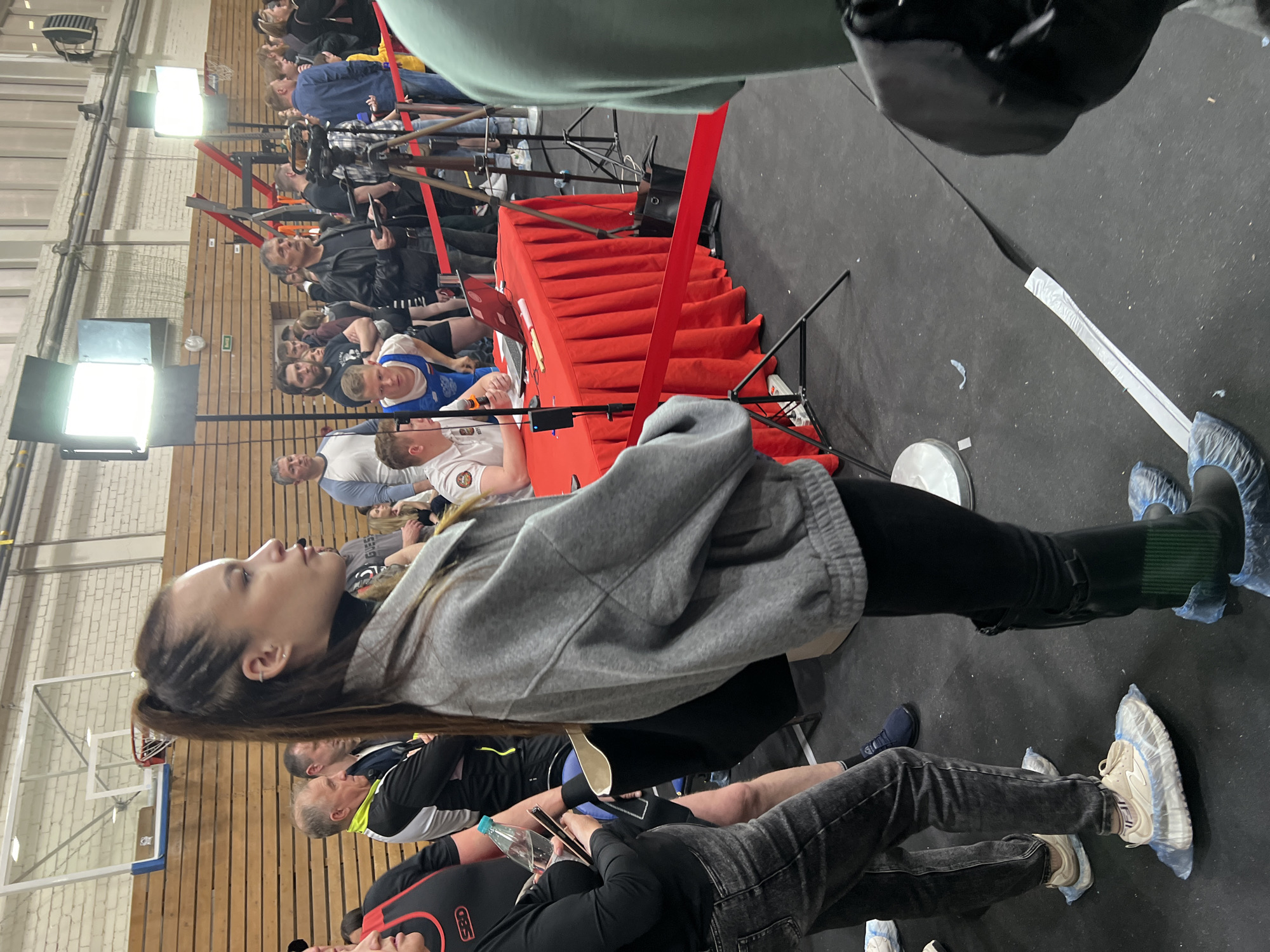 The image depicts an indoor event, possibly a sports competition, in what appears to be a gymnasium-like setting with a wooden wall and a basketball hoop visible in the background. A woman in a gray hooded sweatshirt and black pants is prominently featured, with her face visible. A red-covered table with a red ribbon barrier is set up in the middle of the scene. Several people are sitting or standing in the background. The footwear of some individuals has blue protective coverings. The lighting appears to be a combination of natural light from windows and artificial lights. The scene does not contain definitive elements that would confirm that it is in Saint Petersburg, Russia.