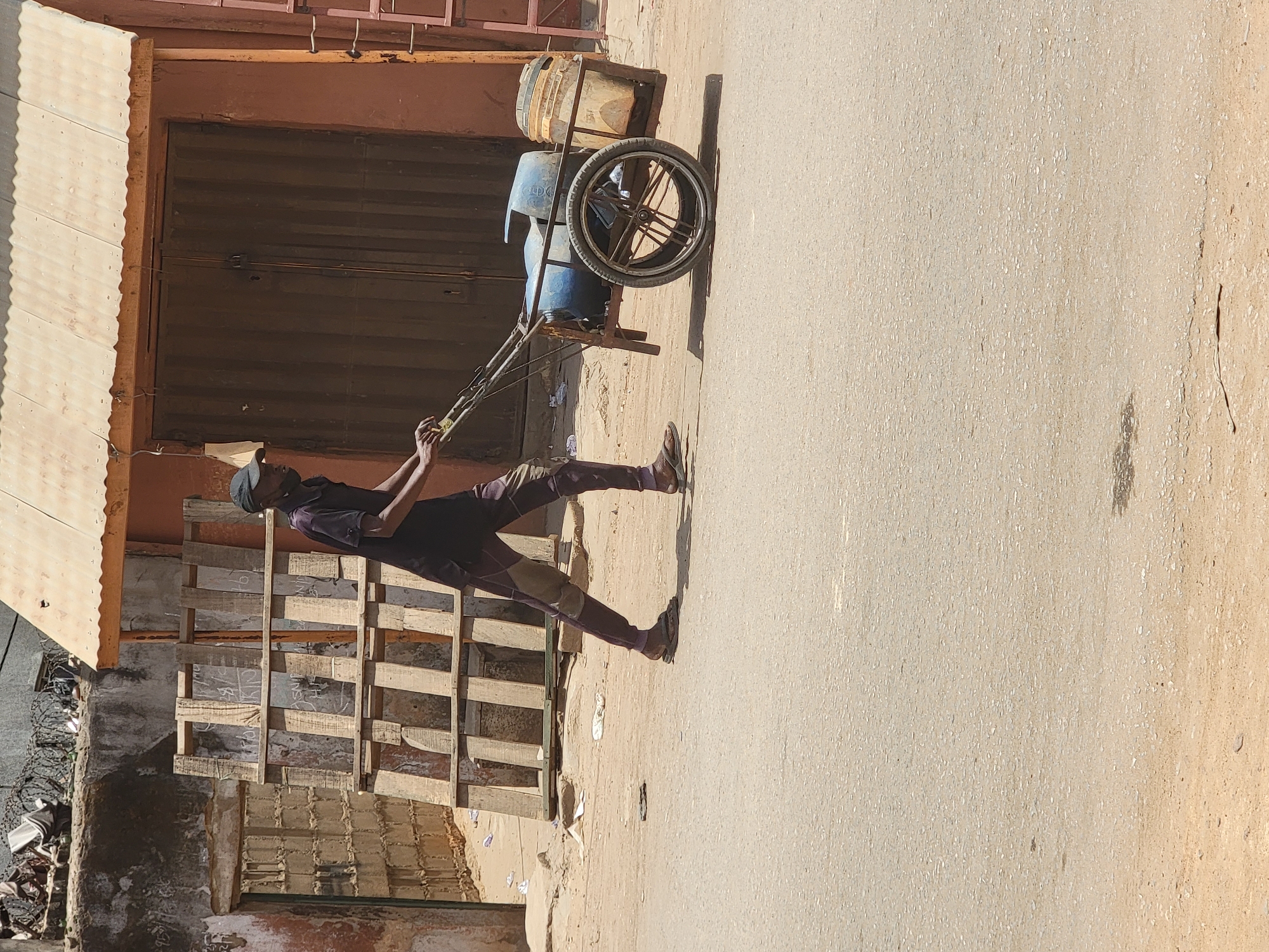 A man wearing a dark shirt and pants is pulling a two-wheeled cart loaded with blue plastic barrels and a white bucket in the city of Maiburiji, Nigeria. The cart is constructed of metal with spoked wheels and a pull rope. The man is barefoot and wearing a cap. He is pulling the cart along a dusty street. To the left of the man is a building facade with a wooden awning, closed metal doors, wooden pallets, and debris. The street is light brown, and the lighting indicates a bright, sunny day.