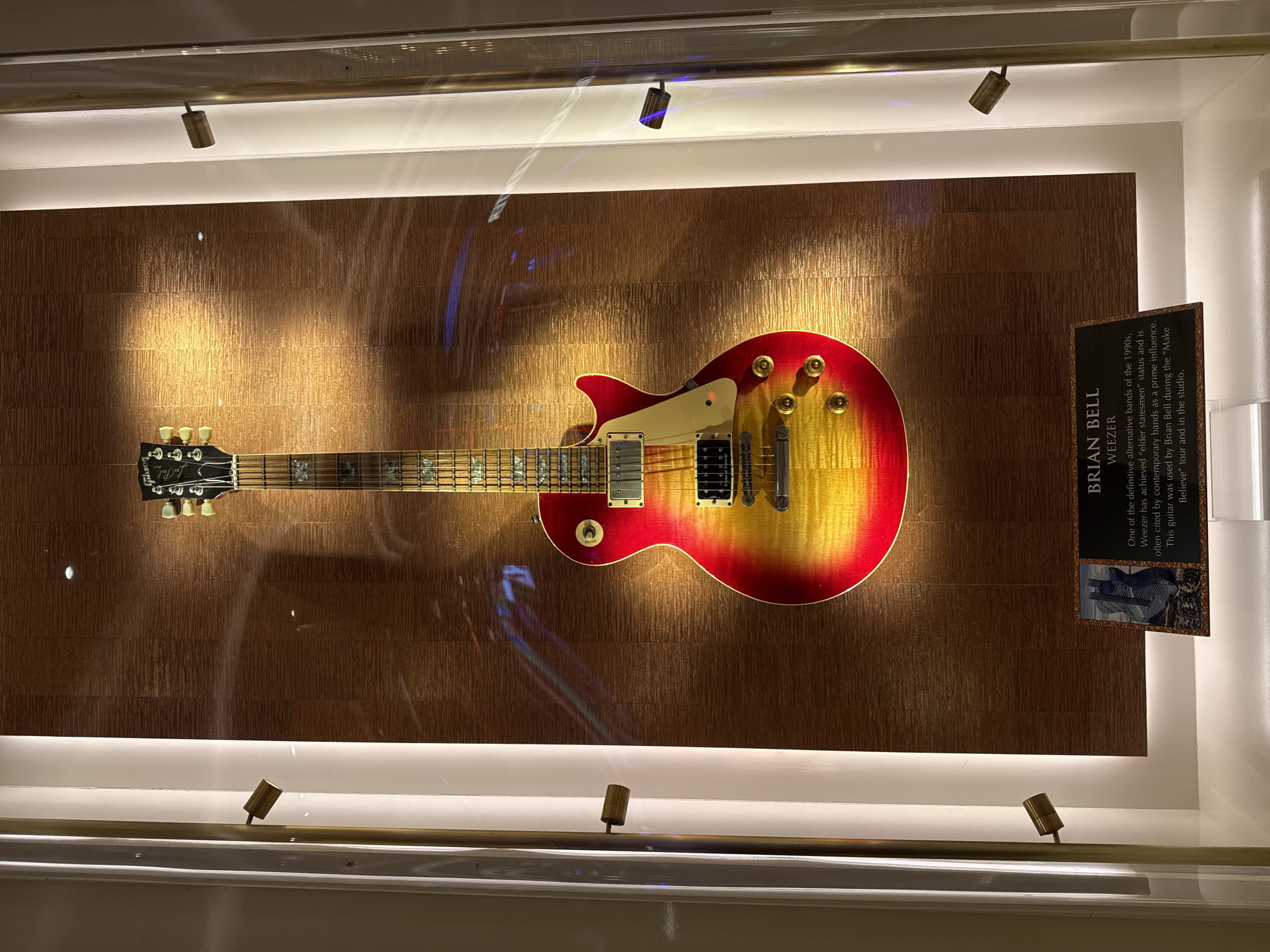 The image captures Brian Bell of Weezer's guitar displayed in a case. The guitar, a Gibson Les Paul with a red and yellow sunburst finish, is mounted against a textured, metallic gold background. Above and below the guitar are small track lights illuminating the display. To the right of the guitar is a plaque with information about Brian Bell and Weezer. The text on the plaque mentions Weezer's influence as a 1990s band and states that the guitar was used during the "Make Believe" tour and in the studio. The photograph was taken at the city of Hollywood Seminole Reservation, United States of America.
