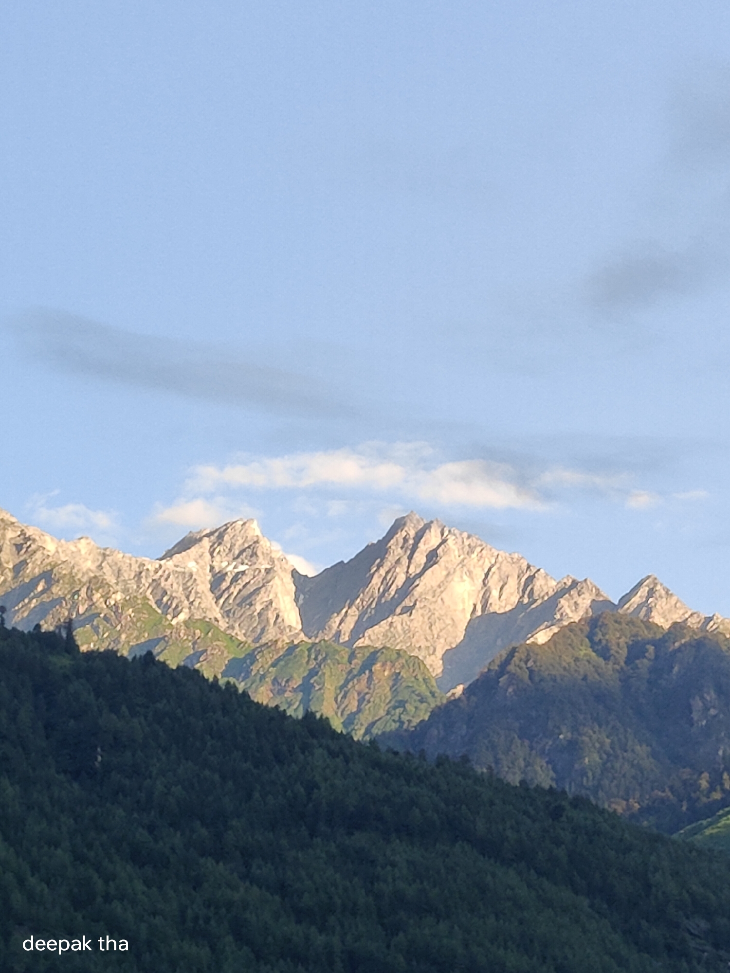 The image shows a mountainous landscape. In the foreground, a dense, dark green forest covers the lower slopes of a hill. Behind the forest, a range of jagged, light-colored mountains rises sharply. The peaks are partially illuminated by sunlight, creating strong contrasts and highlighting their rocky texture. Some patches of snow or ice can be seen on the upper slopes. The sky above is a clear, pale blue with a few wispy clouds. The overall scene is one of natural grandeur and serenity. Text overlays at the bottom left read "deepak tha".