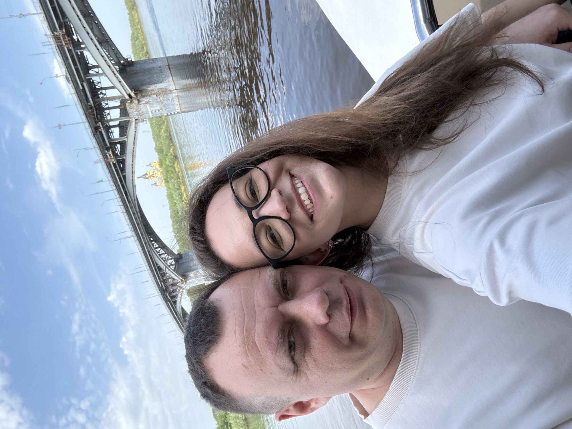 A couple poses for a selfie with the Metromost bridge in Nizhny Novgorod, Russia, in the background. The woman, wearing glasses, smiles widely, while the man gives a more subdued expression. Both are wearing white shirts. Behind them, the bridge spans a body of water with tree-lined banks. The sky is partly cloudy.