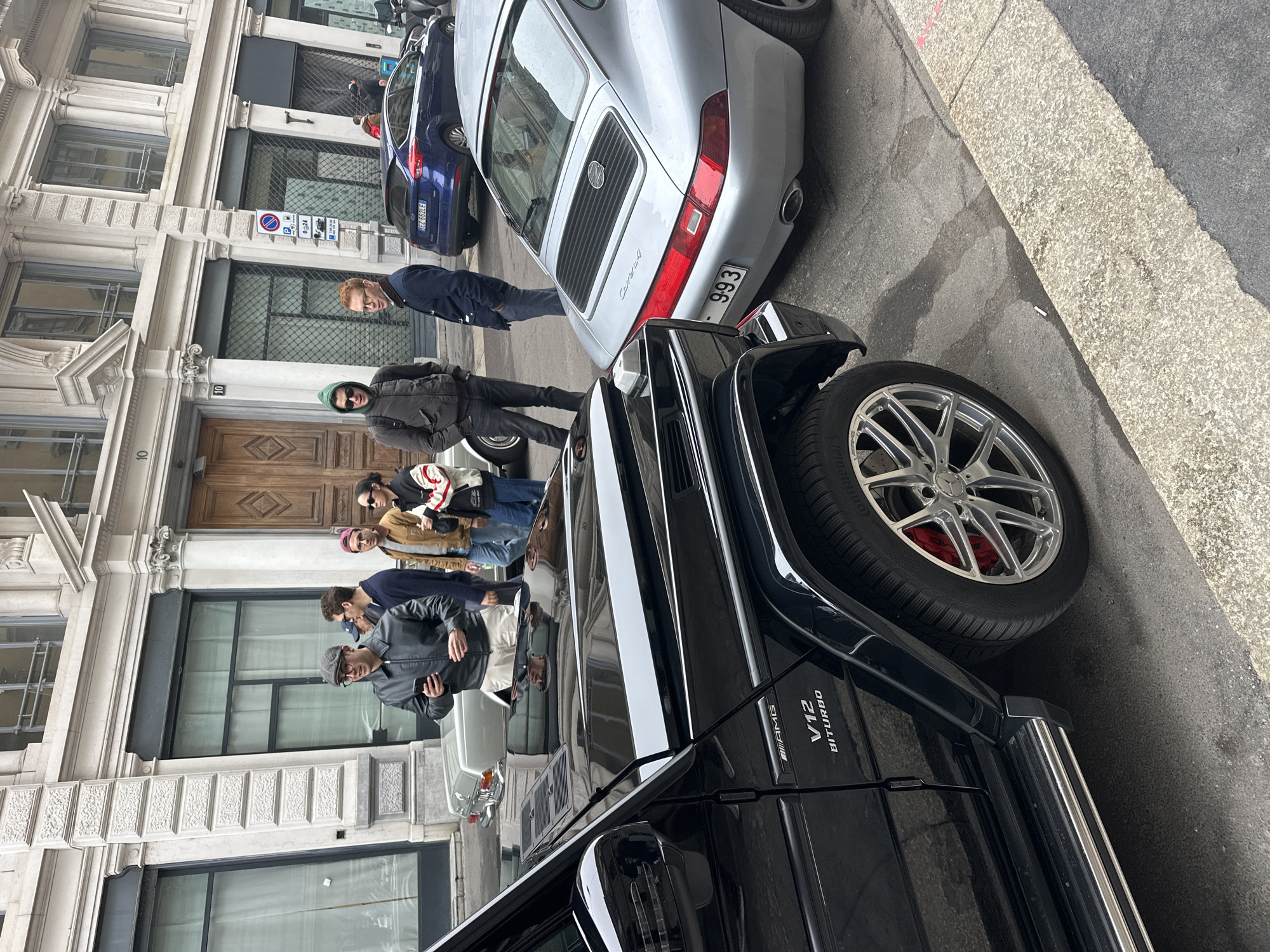 An urban street scene in Milan, Italy, depicts a group of six pedestrians and several vehicles. In the immediate foreground, a black Mercedes-Benz G-Class AMG V12 Biturbo SUV is partially visible on the left, displaying chrome multi-spoke wheels with red brake calipers and "V12 BITURBO" badging. To its right, the rear section of a silver Porsche 911 Carrera 4 (993 generation) coupe is parked, featuring a rear license plate partially displaying "993".
Six individuals are positioned on the asphalt and sidewalk area. From left to right: a man in a grey flat cap and black leather jacket holds a smartphone, looking down; a man in a green hooded jacket and dark sunglasses stands with hands in his pockets; a man with reddish hair and glasses wears a dark blue jacket, looking towards the right; a man in a brown jacket and blue jeans, with a red baseball cap partially visible, looks to the left; and a woman with dark sunglasses, wearing a black and white jacket with red detailing, holds a small bag, looking right. Another man is partially obscured behind the first man in the green hoodie.
Further down the street, a light grey vintage sedan is partially visible. A blue Ford Focus hatchback (license plate EZ 652LE) is parked beyond the pedestrians, followed by several scooters. The background features a multi-story building with a light-colored stone facade, exhibiting classical architectural elements such as columns, cornices, and decorative window frames. Two prominent wooden entrance doors are marked with the number "10" above them. Ground-floor windows are visible, some with interior grates. A blue circular street sign indicating parking restrictions and information is visible on a building column. The ground consists of asphalt road and a stone-paved sidewalk. A white cigarette butt lies on the asphalt near the sidewalk.