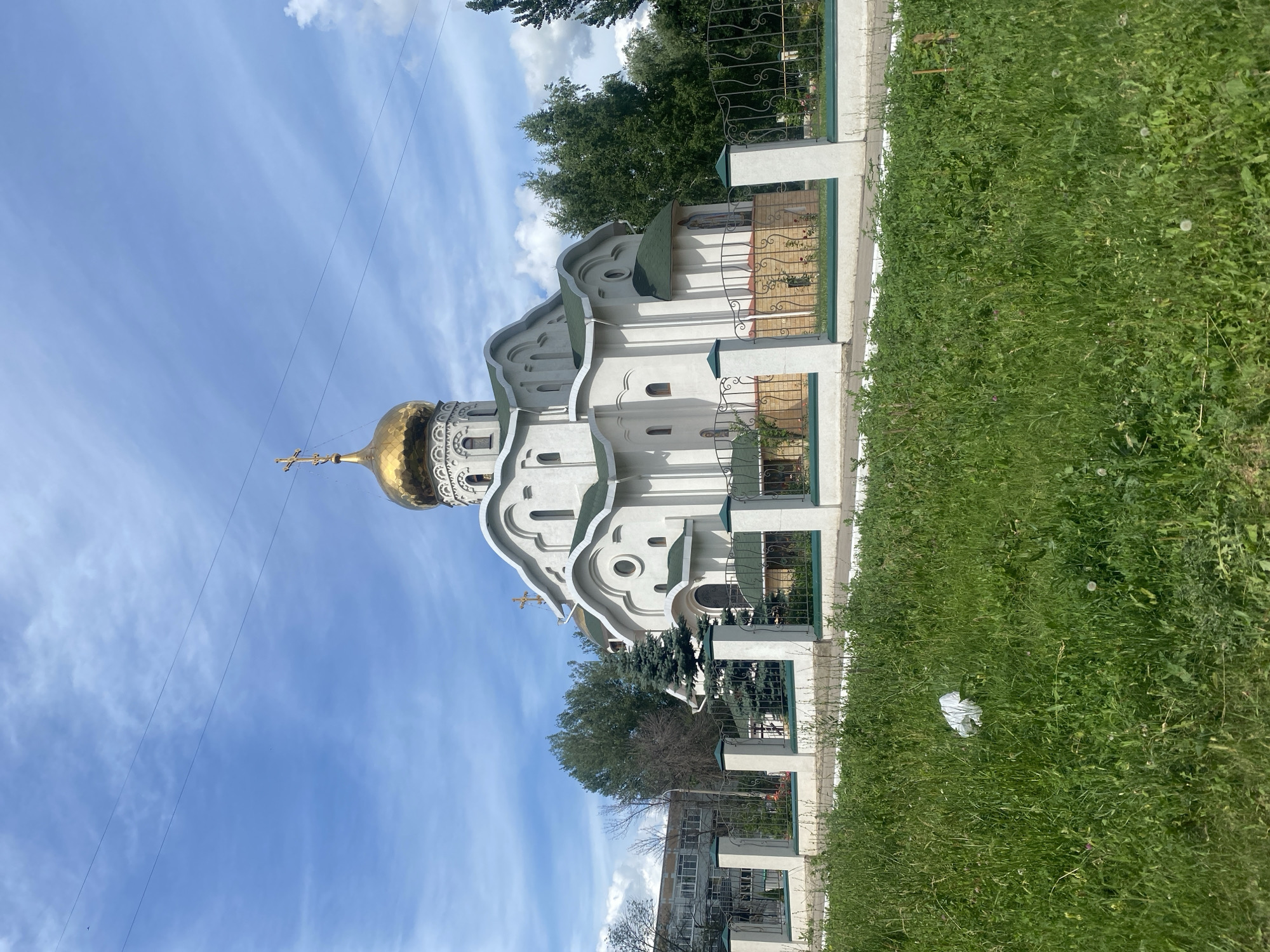 A white Orthodox church with a golden dome topped by a cross stands on a sunny day with a blue sky and scattered clouds. The church has green roofs and several arched windows. A white metal fence with decorative elements separates the church grounds from a grassy area in the foreground. In the background, trees and a multi-story building are visible. The scene is likely a public space in Samara, Russia, given the architectural style and the presence of an Orthodox church.