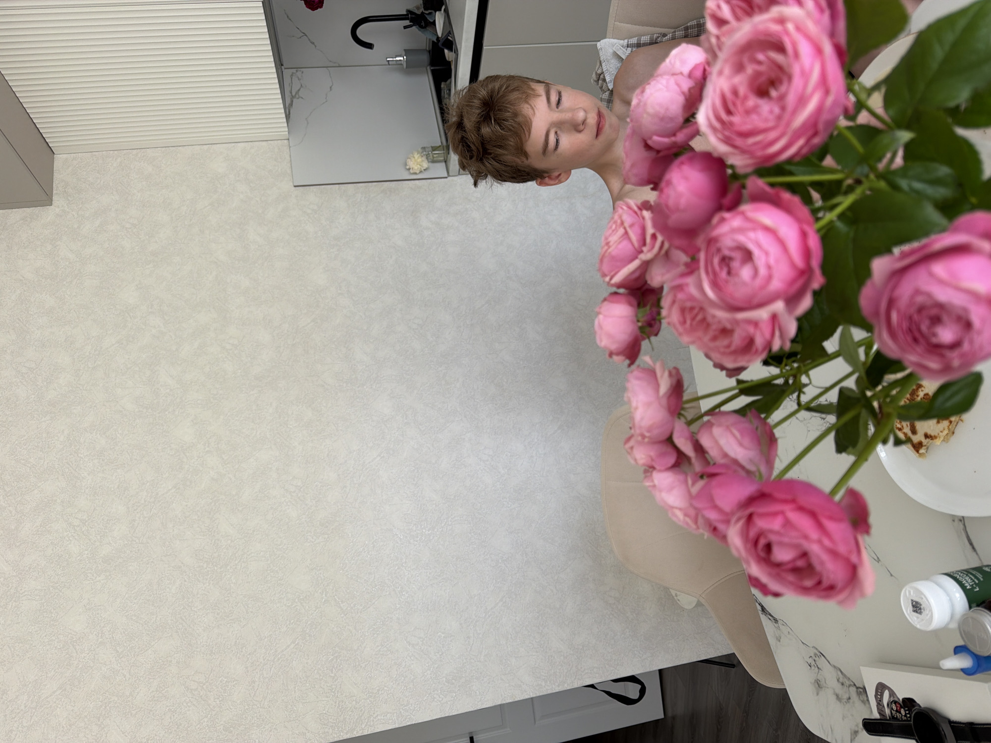 In a brightly lit, modern kitchen or dining area, a young male with short, light brown hair is seated at a table, looking downwards. He is shirtless, with a light-colored plaid garment draped over his shoulders. In the foreground, a bouquet of vibrant pink roses is arranged in a vase, partially obscuring the table and the lower portion of the individual. The table itself is round with a marble-patterned surface. On the table, several items are visible: a white box, a watch, a bottle with a label, and a plate with a folded pastry, possibly a crepe or pancake. The background features a textured light-colored wall and a section of a white kitchen counter with a black faucet and a bottle of soap dispenser. The overall setting suggests a domestic interior.