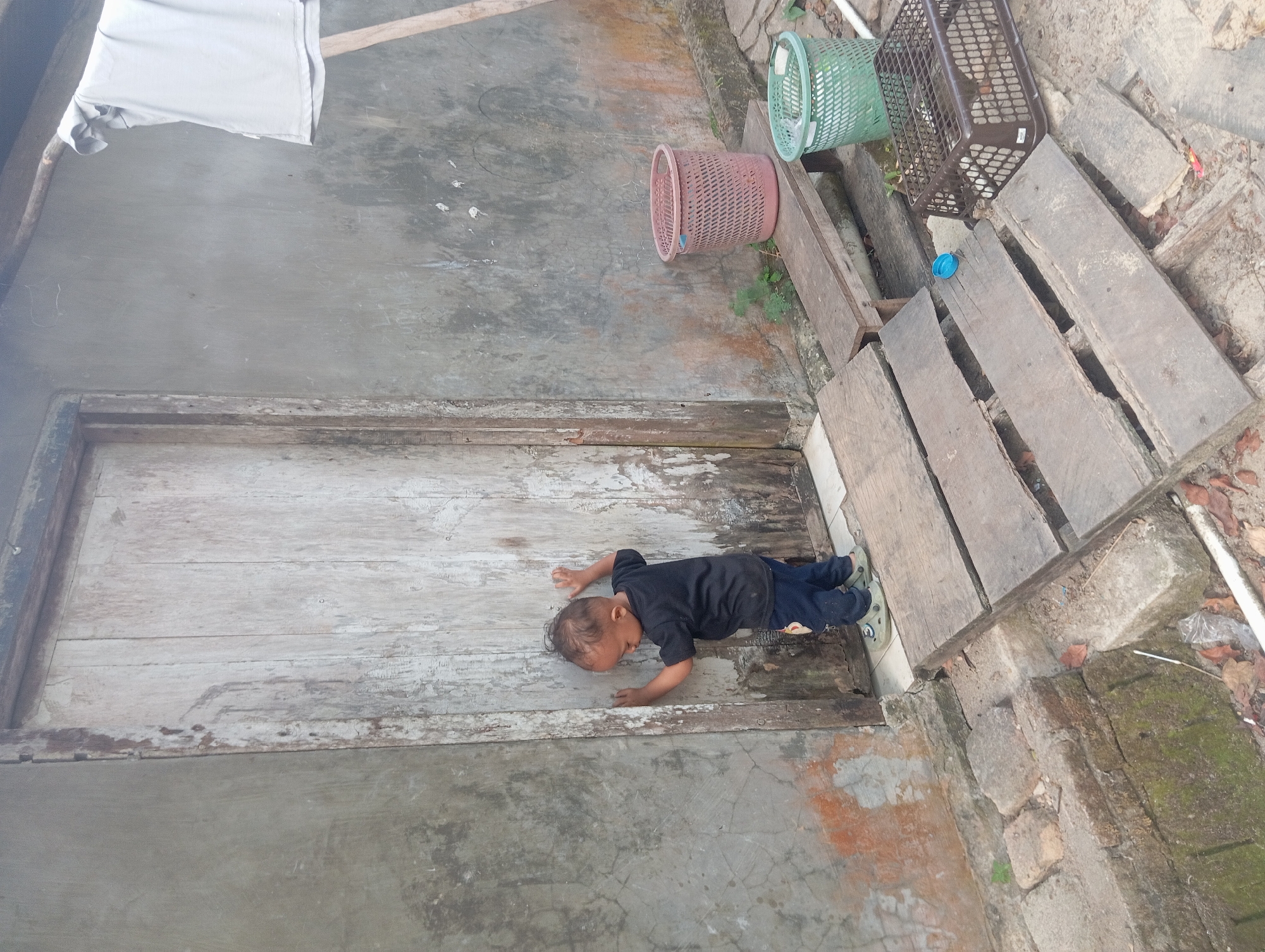 A young child, appearing to be approximately two years old, stands with their back to the camera, leaning against a weathered wooden door. The child is wearing a black t-shirt and dark blue pants. Their head is tilted downwards, and their hands are pressed against the door's surface. The door itself is made of vertical wooden planks, showing signs of age and wear with peeling paint and exposed grain.
The door is set into a rough, grey concrete wall, which is stained and textured. To the right of the door, a wooden bench supports two baskets: a pink, woven basket and a green, plastic basket. Further to the right, a brown, plastic crate with a grid pattern sits on the ground.
In front of the door, a makeshift ramp constructed from wooden planks leads up to the door. The planks are irregularly spaced and appear weathered. A small blue object, possibly a bottle cap, rests on one of the planks.
The ground around the ramp and baskets is covered in dirt, leaves, and small stones. To the far right, a piece of light-colored fabric, possibly clothing, is draped over a wooden pole, suggesting an outdoor laundry area. The overall environment appears to be an outdoor space attached to a simple dwelling, likely in a rural or semi-urban setting. The scene suggests a moment of childhood exploration or curiosity.