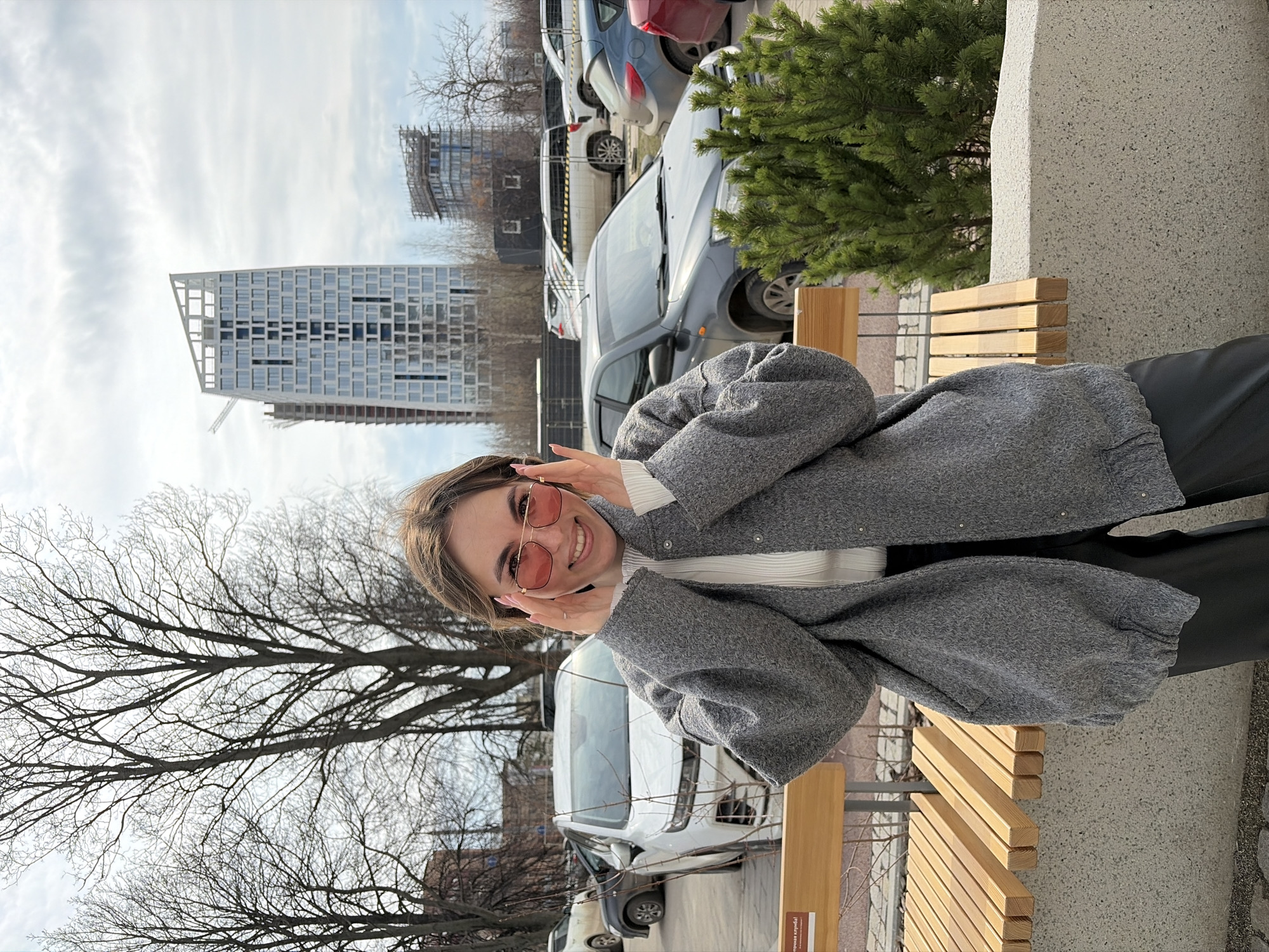 The image shows a woman posing on a wooden bench outdoors. She has light brown hair, wears pink sunglasses, a white sweater, and a gray coat. She is smiling and holding her hands near her face. In the background, there are leafless trees, parked cars, a modern high-rise building under construction, and a smaller building also under construction. A small evergreen tree sits beside the bench. Based on the architecture and visible signs, the location is Izhevsk, Russia. The scene appears to be an urban area on a cloudy day.