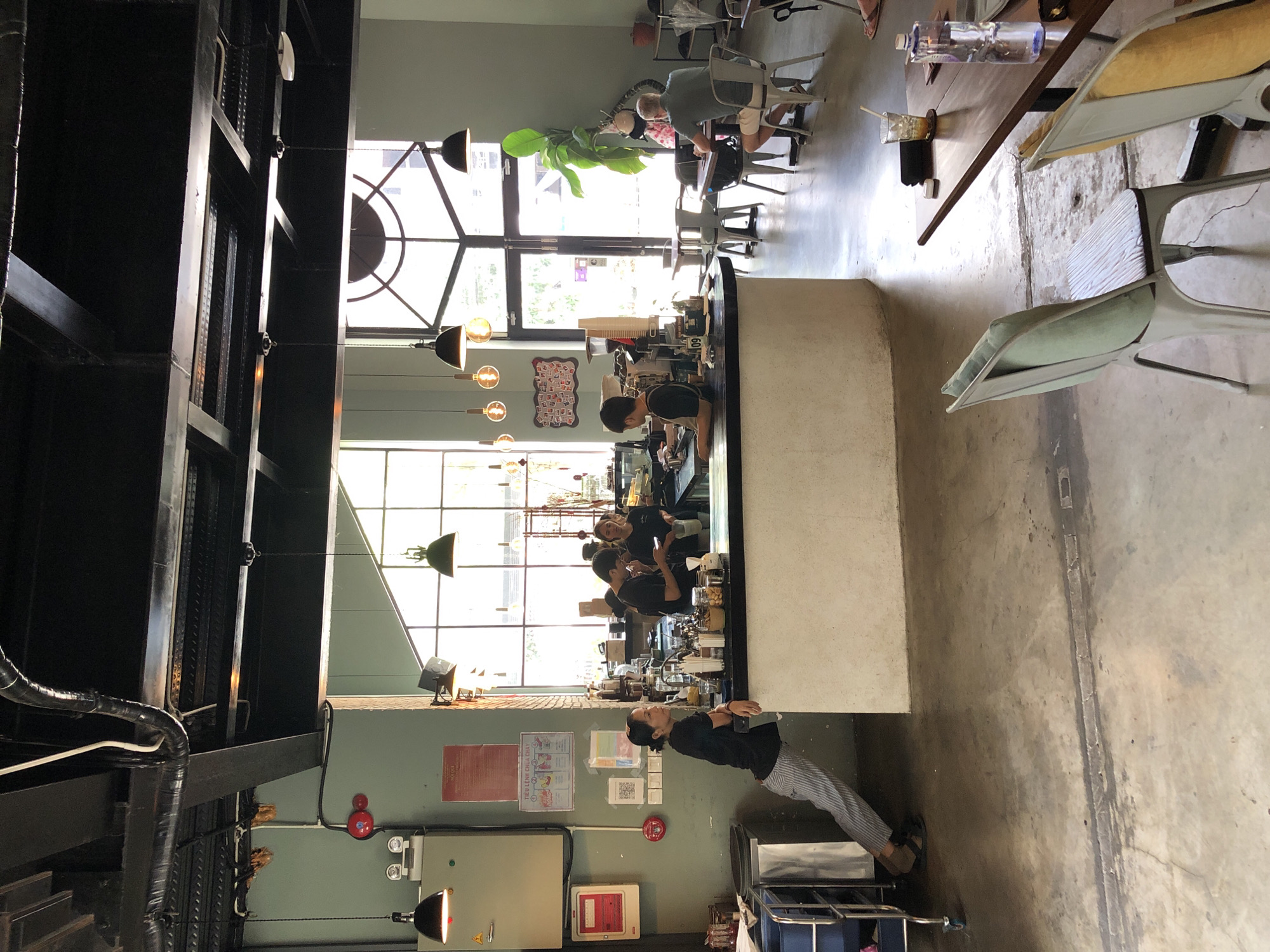 The image shows the interior of a cafe in Da Nang, Vietnam. The cafe has a modern industrial aesthetic with exposed ductwork and concrete floors. Several people are present, including baristas working behind a counter and customers sitting at tables. One woman in striped pants is leaning on the counter, looking towards the back of the cafe. A man in a green shirt is seated at a table, facing away from the camera. Another man in a black t-shirt is working behind the counter. Natural light streams in from large windows at the back of the cafe, illuminating the space. The cafe appears to be open for business, with food and drinks visible on the counter and tables.
