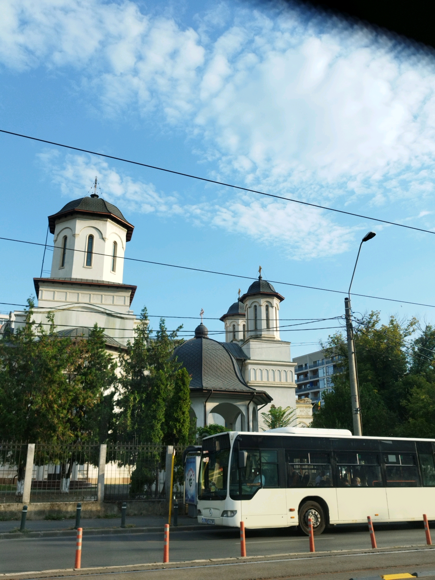 The image captures a street scene in Bucharest, Romania, featuring a white bus in motion on the right side of the frame. To the left, a white church with a dark dome and prominent windows stands behind a black metal fence and lush green trees. The sky above is a clear blue, dotted with scattered white clouds. A modern apartment building is visible in the background, to the right of the church. The street is paved, with traffic cones and bollards delineating the road. The bus displays the license plate "B 003 TTK". Passengers are visible through the bus windows.