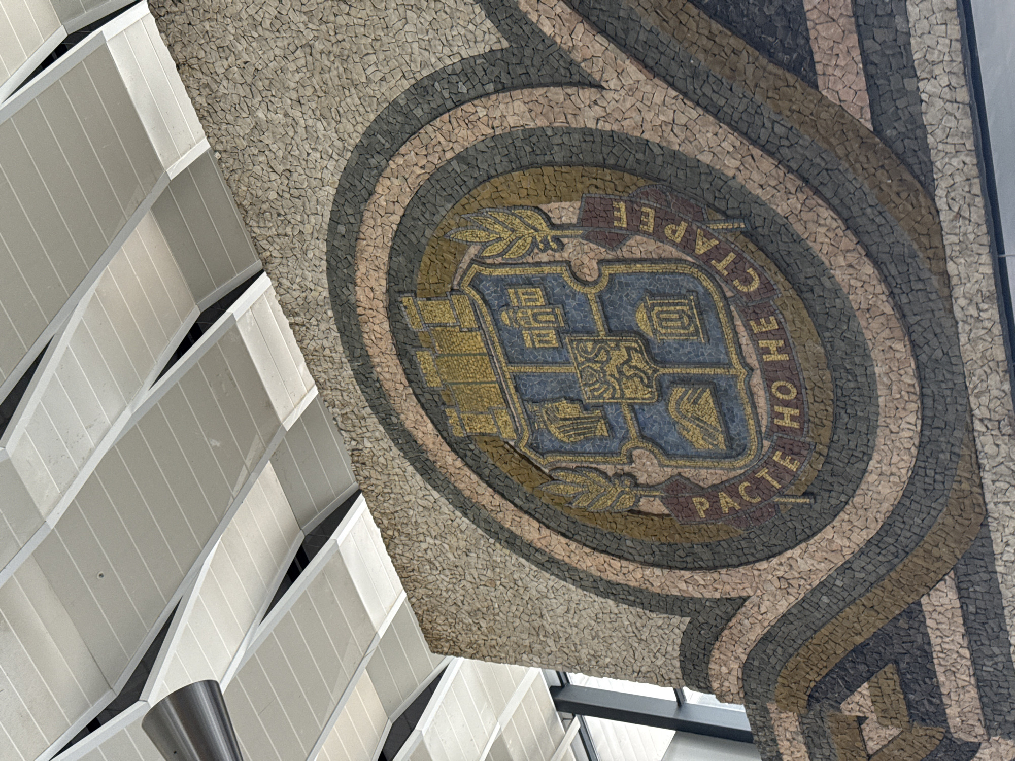The image captures a mosaic ceiling decoration within the Palace of Justice in Sofia, Bulgaria. The mosaic features the Coat of Arms of Sofia, encircled by the inscription "РАСТЕ, НО НЕ СТАРЕЕ", which translates to "It grows, but does not age." The coat of arms is composed of gold and blue tesserae. The outer edges of the circular mosaic are layered with tesserae of brown, gray, and off-white hues. In the background, a portion of the building's architecture is visible, displaying a sequence of white angled panels.