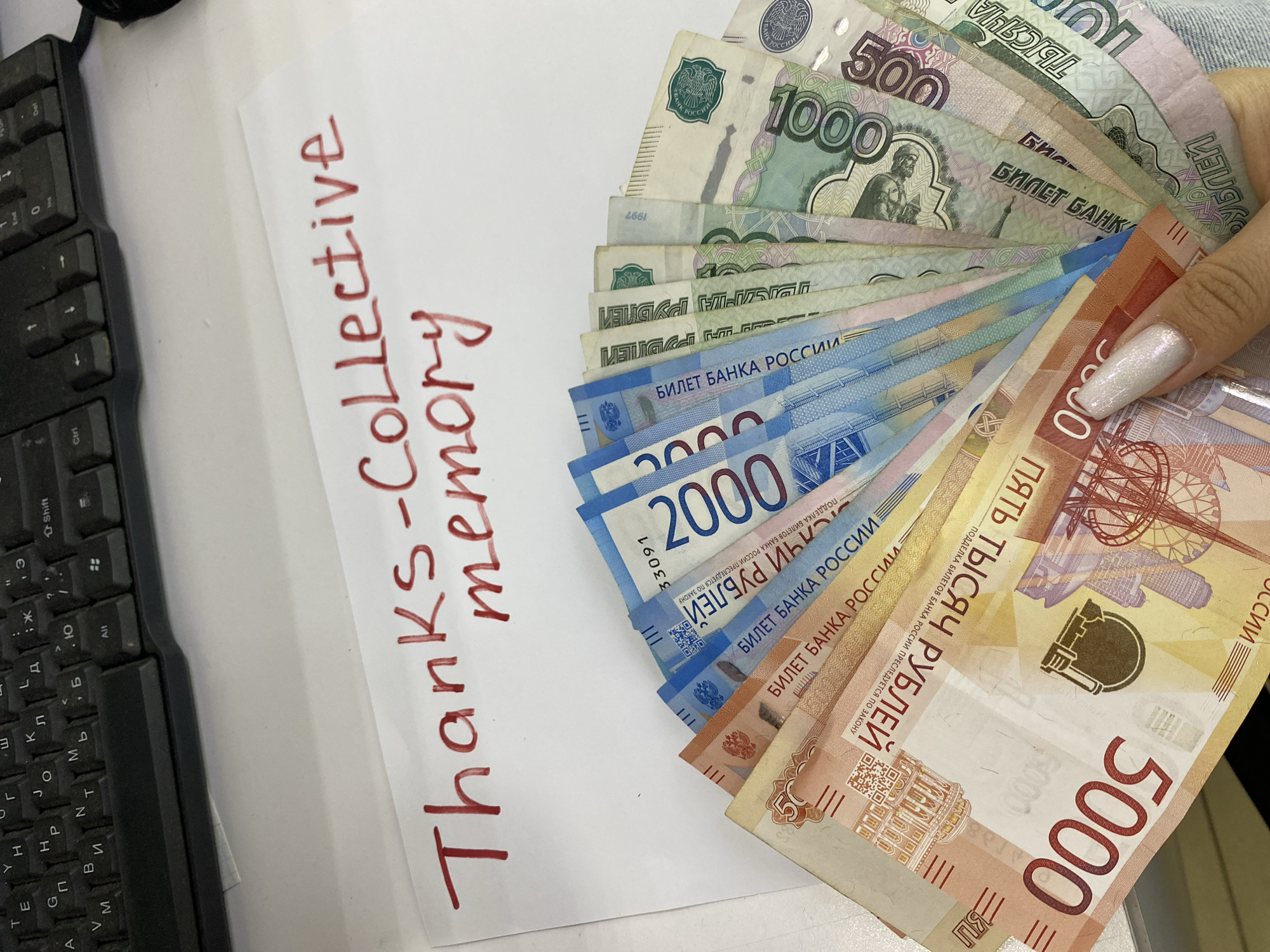 The image depicts a white desk with a black computer keyboard on the left. A white piece of paper with the handwritten phrase "Thanks-Collective memory" in red ink sits on the desk. To the right, a hand holds a fan of Russian rubles in various denominations: 5000, 2000, 1000, and 500. The banknotes are predominantly in the colors of pink/orange, blue, green, and purple respectively. The setting is unidentified, and the location within the city of Chaikovsky, Russia, cannot be confirmed from the image.