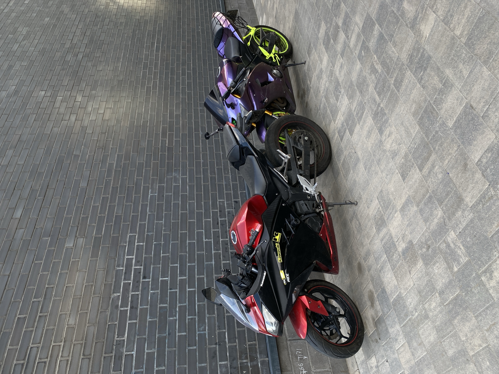 The image displays two motorcycles parked side-by-side against a dark gray brick wall in an outdoor urban setting in Saint Petersburg, Russia.
The motorcycle on the left is a Kawasaki sportbike, predominantly red and black. Its front fairing, fuel tank, and lower fairing panels are red, while the side panels and seat are black. "Kawasaki" branding is visible on the side fairing and tank, alongside a yellow "K" logo and "MIX" text. It features black multi-spoke wheels with a thin red stripe on the rims and is supported by a black kickstand.
The motorcycle on the right is also a sportbike, featuring an iridescent purple/violet paint scheme that appears to shift color. Its frame and rear swingarm are a bright neon yellow/lime green, matching the color of its multi-spoke wheels. The seat is black, and a black metal cargo rack is mounted on the rear fender. This motorcycle is also supported by a black kickstand.
The background consists of a dark gray brick wall with lighter gray mortar lines, laid in a running bond pattern. The lower portion of the wall, below the main brickwork, is a lighter concrete base with some faint white markings, including what appears to be "$66 J171". The ground surface is paved with interlocking concrete pavers in varying shades of gray, arranged in a herringbone pattern. The scene is illuminated by natural daylight. No individuals are present in the image.
