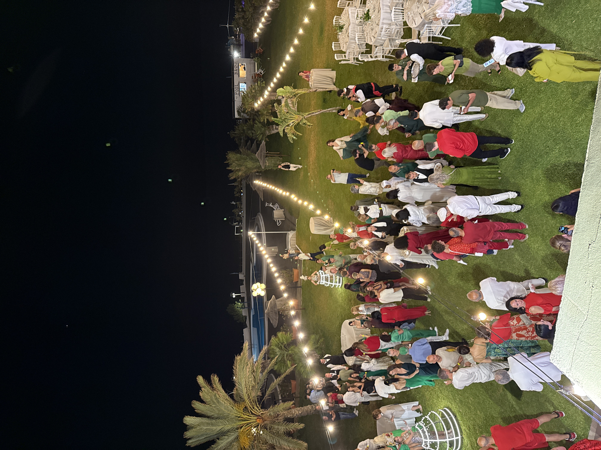 The image shows an outdoor evening event in Isola delle Femmine, Italy. People are gathered on a green lawn, illuminated by string lights. A pool and palm trees are visible in the background. The crowd appears to be mostly adults, wearing varied clothing, including shades of red, green, and white. Some are standing, while others seem to be seated or lying down. Tables with white chairs are present on the lawn.