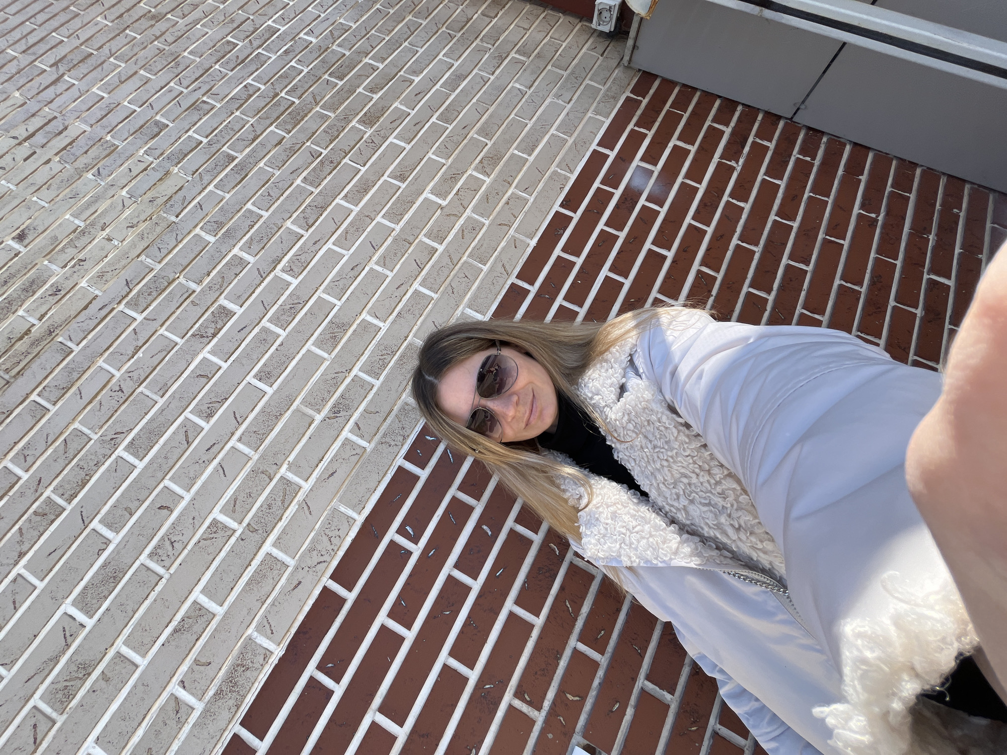 A selfie shows a woman with blonde hair and sunglasses wearing a white jacket standing on a brick walkway. The brick walkway is composed of tan bricks and red bricks. The walkway appears to be adjacent to a building. The location is possibly in Chaikovsky, Russia.
