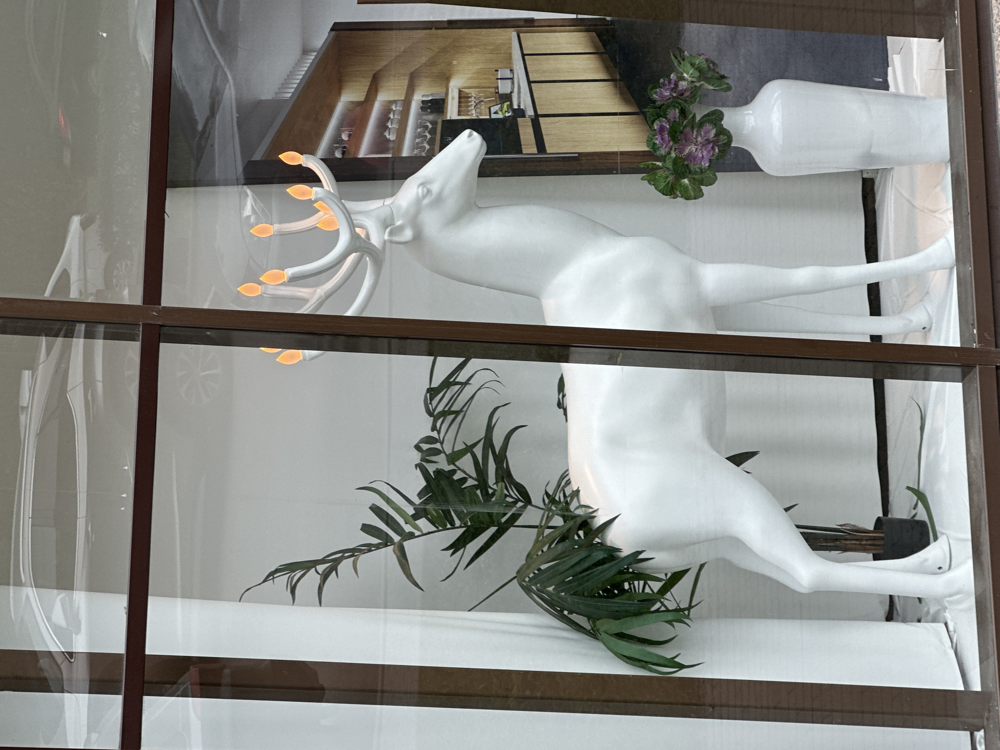 This is a medium shot, eye-level photograph of a white deer statue with illuminated antlers, displayed in a store window. The deer is positioned centrally, facing to the right, with its head slightly lowered. Its antlers are shaped like branches and tipped with glowing orange lights, resembling flames or candles. To the left of the deer, a tall green plant in a black pot leans towards the statue. In front of the deer's hindquarters and to the right of the statue, a tall white vase holds a cluster of purple and green leafy decorative plants. The background of the window display features a printed image of a modern kitchen with wooden cabinets and shelves stocked with bottles and what appear to be decorative items. Reflections on the glass of the window include a silver car and parts of the surrounding environment. The display is framed by dark brown window mullions. The floor of the display area is covered in white fabric.
