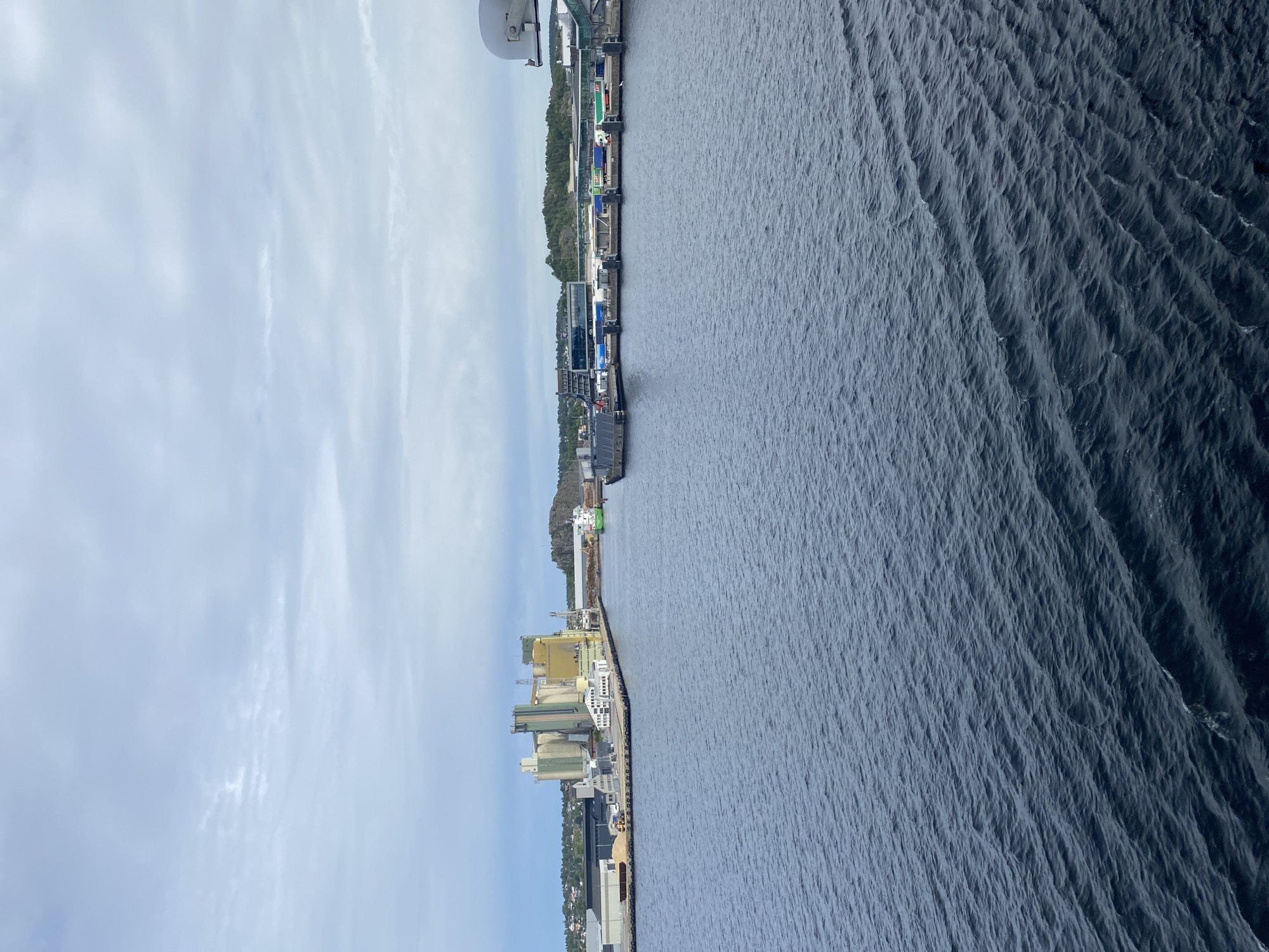 The image shows a port in Larvik, Norway, with a body of water in the foreground and industrial buildings along the coastline in the background. The water is dark blue and appears to have small waves. The buildings are a mix of modern and industrial structures, with some appearing to be grain silos or storage facilities. There are also some cranes visible, suggesting ongoing activity at the port. The sky is overcast with some clouds. The time of day appears to be daytime, given the lighting. The overall scene is industrial and functional, typical of a working port. There is no visible text in the image.