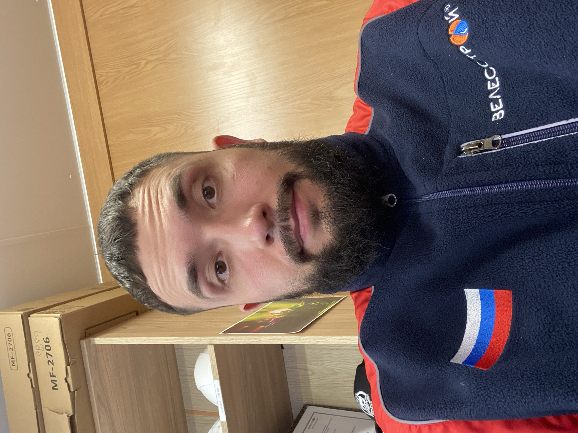 A selfie shot shows a bearded man wearing a blue and red jacket with a Russian flag patch. "BEAECOTP" is embroidered in white near the shoulder with a logo above. The man is indoors, standing in front of wooden shelves, which hold boxes labeled "MF-2706," a white hard hat, a framed certificate, and a picture of what appears to be fireworks. There is no explicit indication of Luzhitsy, Russia in the image.
