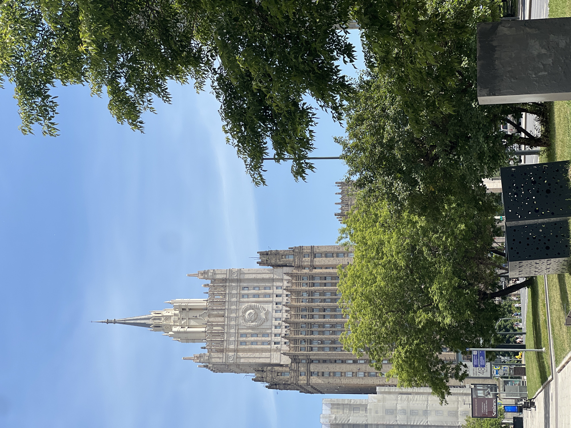 The image displays the Ministry of Foreign Affairs building in Moscow, Russia, under a clear blue sky with minimal cloud cover. The prominent central subject is a tall, beige-colored skyscraper, characterized by its Stalinist Empire architectural style, featuring a distinctive central spire that tapers to a fine point. A large circular emblem, resembling the Soviet Union's coat of arms, is prominently situated on the upper facade of the main building block. The building exhibits multiple vertically stacked sections with numerous rectangular windows, many of which have visible external air conditioning units.
In the foreground, a vibrant green grassy median is populated with several deciduous trees, their lush foliage partially obscuring the lower portions of the building. To the left, a paved sidewalk runs parallel to a street. Visible along the sidewalk are various street signs, including one with the text "Sminex" and another blue sign displaying "48" and Cyrillic script. A multi-story building to the far left is partially enveloped in white protective scaffolding or sheeting. On the grassy median, two dark, abstract sculptural objects are present; one is a rectangular block with numerous circular perforations, and the other is a solid, dark rectangular prism. Beyond the trees and grassy area, a street is discernible, with several small figures appearing to be pedestrians walking in the distance. The scene is illuminated by bright, natural daylight.