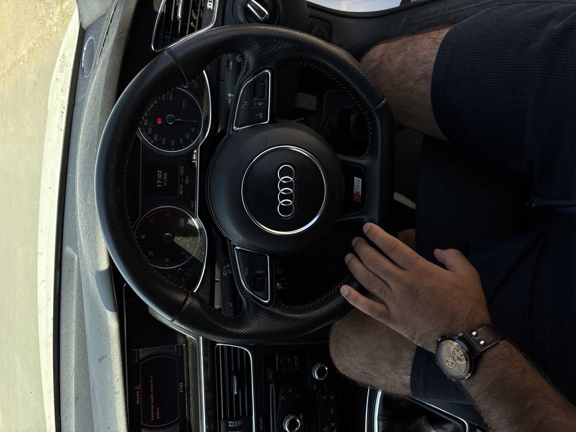 The image shows the interior of a car, specifically the dashboard and steering wheel, as seen from the driver's perspective. The driver's hands are visible on the steering wheel and on their leg. The car's infotainment system displays "External audio source is active" and the time "17:02". The odometer shows "191110 mi" and the temperature is "+28.5°C". The car appears to be an Audi, indicated by the logo on the steering wheel and the "S line" badge. The scene is consistent with a person sitting in the driver's seat of a parked car on a sunny day. The location is identified as Dolni Tsibar, Bulgaria.