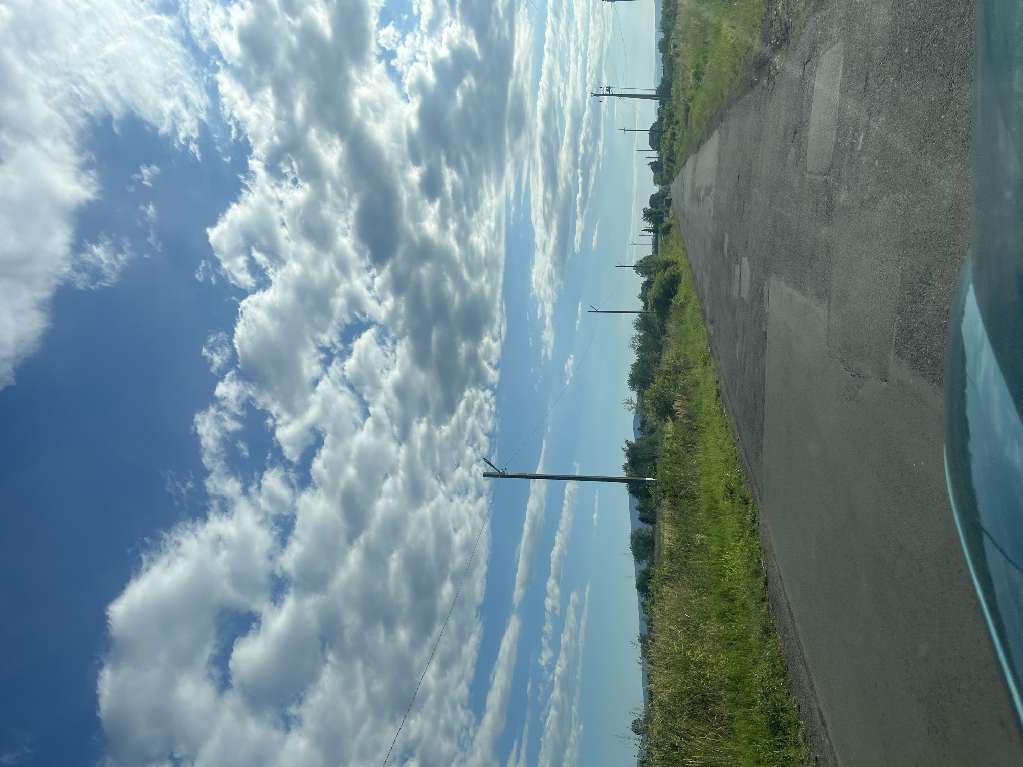 The image captures a view from a vehicle traveling on a paved, somewhat worn rural road. The road extends into the distance, flanked by lush green grassy verges and scattered trees. In the background, rolling hills are visible under a bright blue sky dotted with puffy white clouds. Several utility poles with overhead wires are positioned along the roadside, indicating the presence of infrastructure. The overall scene suggests a pleasant, sunny day in a rural setting, likely in Storozhnytsia, Ukraine, given the context. No people are visible in the frame.