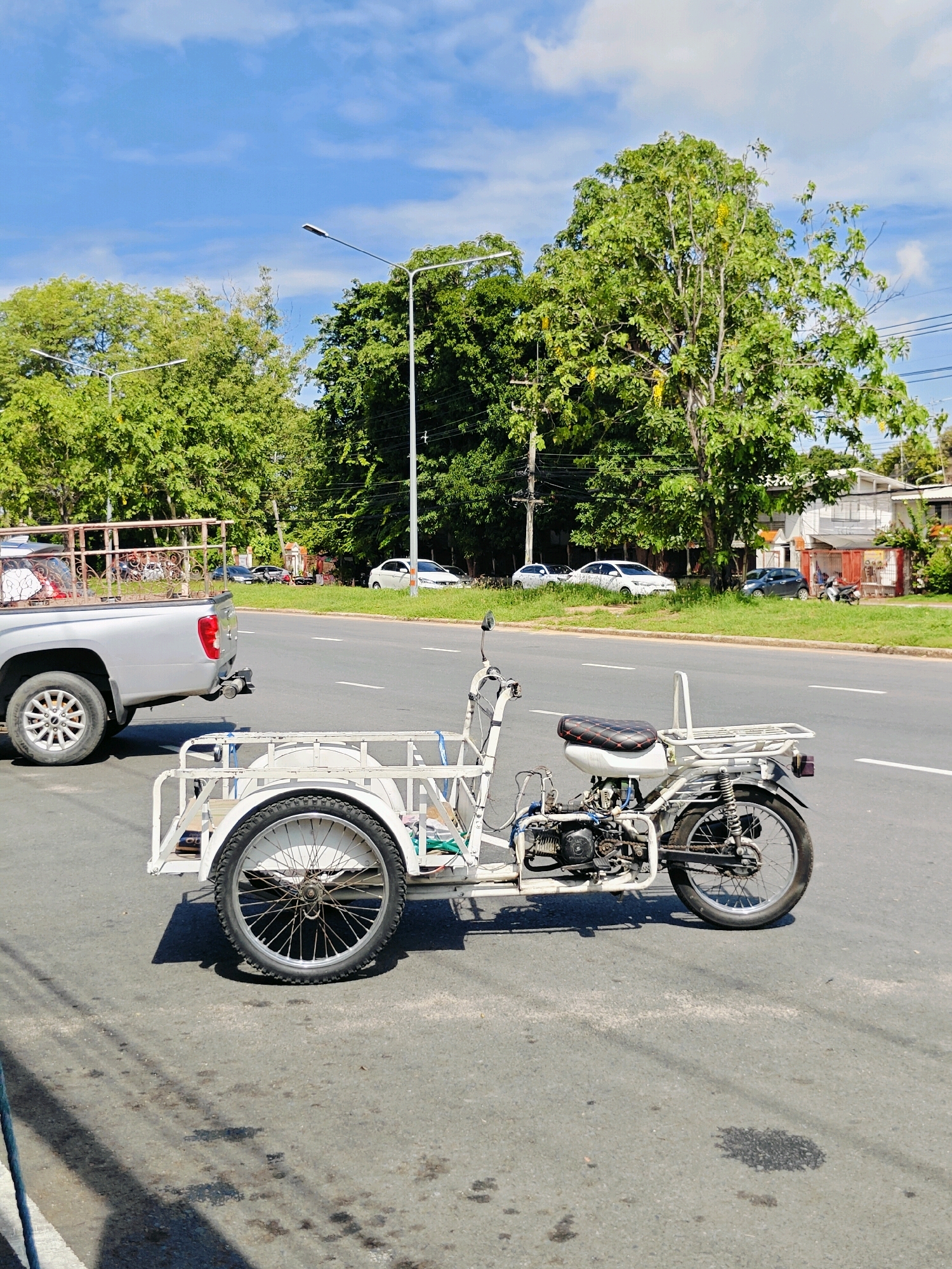 A white, three-wheeled motorized cart with a metal frame and a small engine is parked on the right side of a paved road. The cart has a seat, a rack behind the seat, and a large cargo area in the front. To the left of the cart, a silver pickup truck is also parked on the side of the road. In the background, there are trees, a street light, and several buildings. The sky is blue with some clouds. The scene appears to be in an urban or suburban area, possibly Phra Nakhon Si Ayutthaya City Municipality, Thailand, given the presence of the cart, which is common for local transport and commerce. There are no people visible in the image. The environment suggests a warm, sunny day.