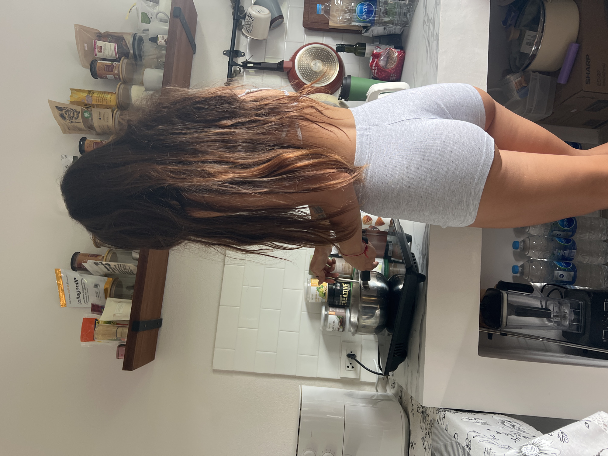 The image depicts a kitchen scene in a residence located in the city of บ้านบางรักษ์, Thailand. A female individual, seen from the back, is standing at a kitchen counter, engaged in cooking or preparing food. She has long, dark brown, wavy hair that falls down her back. She is wearing light gray athletic shorts and a white top, partially obscured by her hair. Her left wrist has a red string bracelet.
She is observed stirring the contents of a stainless steel pot placed on a black induction cooktop. To her left, a white air fryer is visible on the counter, next to a power outlet with a black cord plugged in. The countertop has a white marble-like pattern, and an adjacent table to the left is covered with a white tablecloth featuring a dark floral design.
The wall behind the counter features a white subway tile backsplash. Above the backsplash, two wooden shelves with black metal brackets hold various kitchen and food items. The left shelf contains items such as a "CollagenUP" package, other bags, jars, and a bamboo whisk. The right shelf displays more assorted bags (including "California GOLD Nutrients"), jars with wooden lids, white mugs, and a small dropper bottle.
Behind the cooking area on the counter, several containers are visible, including one labeled "BLACK CHIA" and another prominent one labeled "Micronized CREATINE Powder." Other items on the counter include a green travel mug, a partially visible bag of "BUCKWHEAT," a small bottle of olive oil, and a large "CRYSTAL" brand water bottle. A red frying pan and several mugs hang from a black rail on the wall to the right. A wooden cutting board is also present.
Below the counter, an open cubby on the left houses a black blender with a clear pitcher, alongside multiple "CRYSTAL" brand water bottles. To the right, under the counter, a "SHARP" brand cardboard box and additional kitchen items, including a light-colored pot and a purple cylindrical object, are stored.