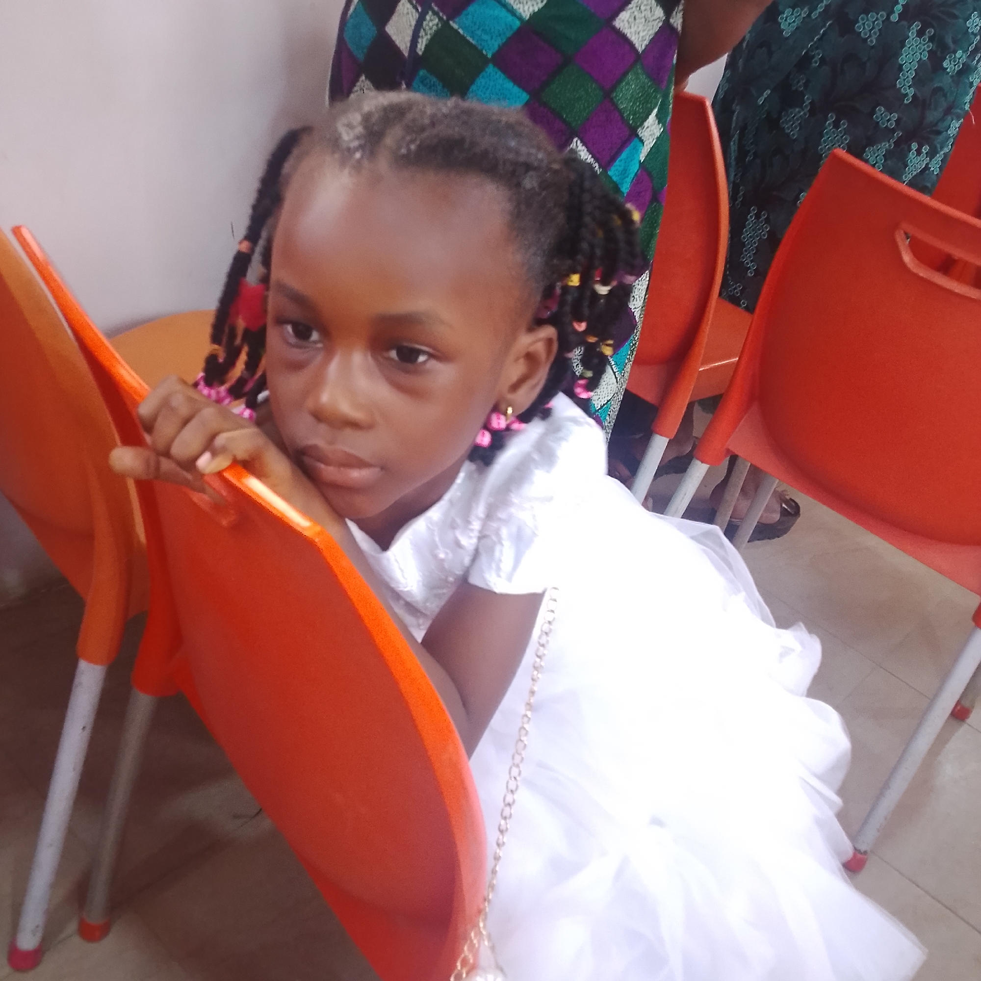 A young Black girl with braided hair is sitting on an orange plastic chair, leaning her head on her hand and looking off to the side. She is wearing a white dress and a gold chain with pearls around her neck. The background shows other orange chairs and people in traditional Nigerian clothing. The scene appears to be indoors, possibly at a gathering or event. The floor is tiled.