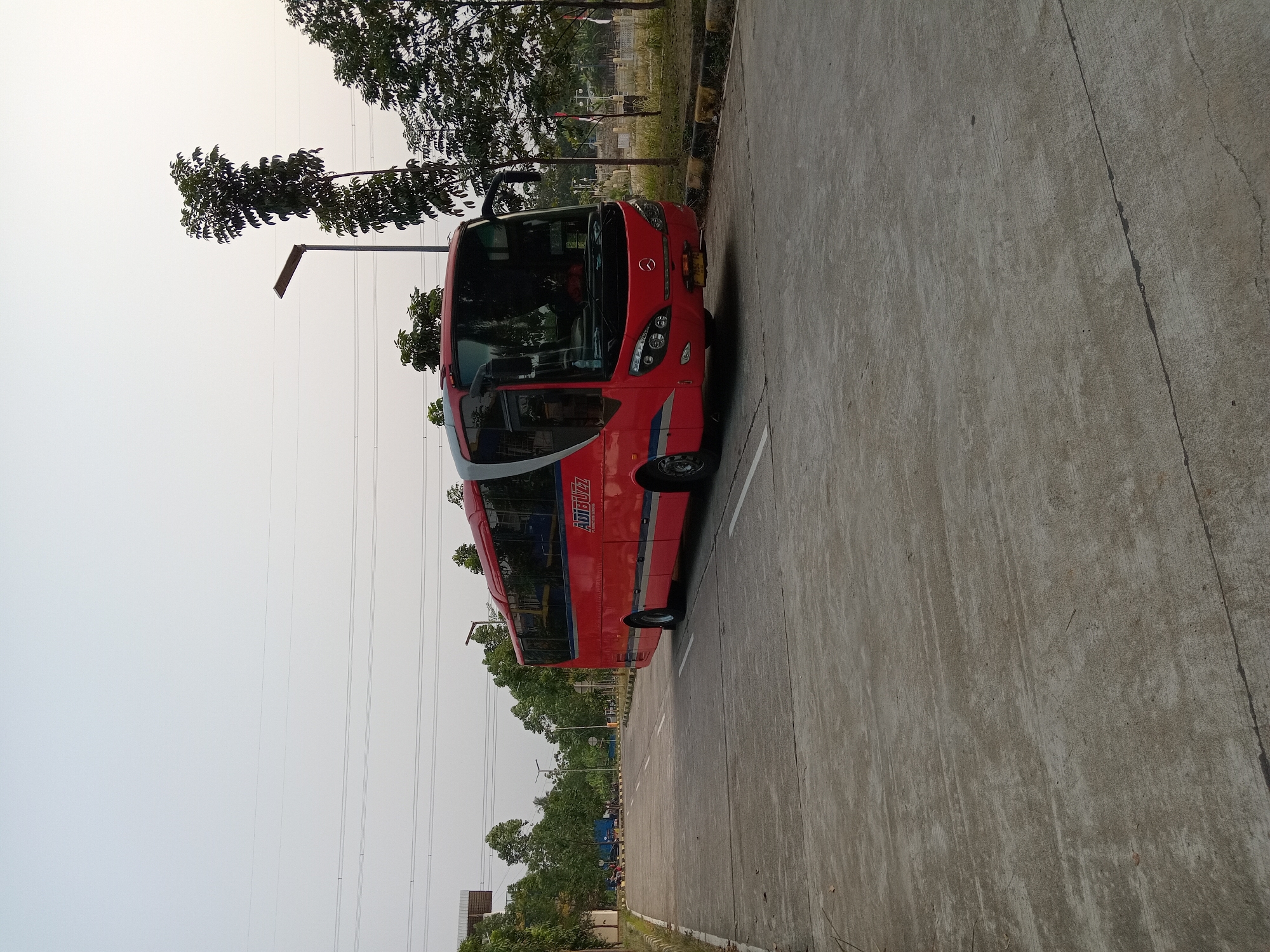 A bright red bus with "ADIBUZZ" text visible on its side is parked on a concrete road. The bus is facing the viewer from the front right. Trees and utility poles are visible in the background, with a slightly overcast sky. The scene appears to be in an industrial or commercial area.