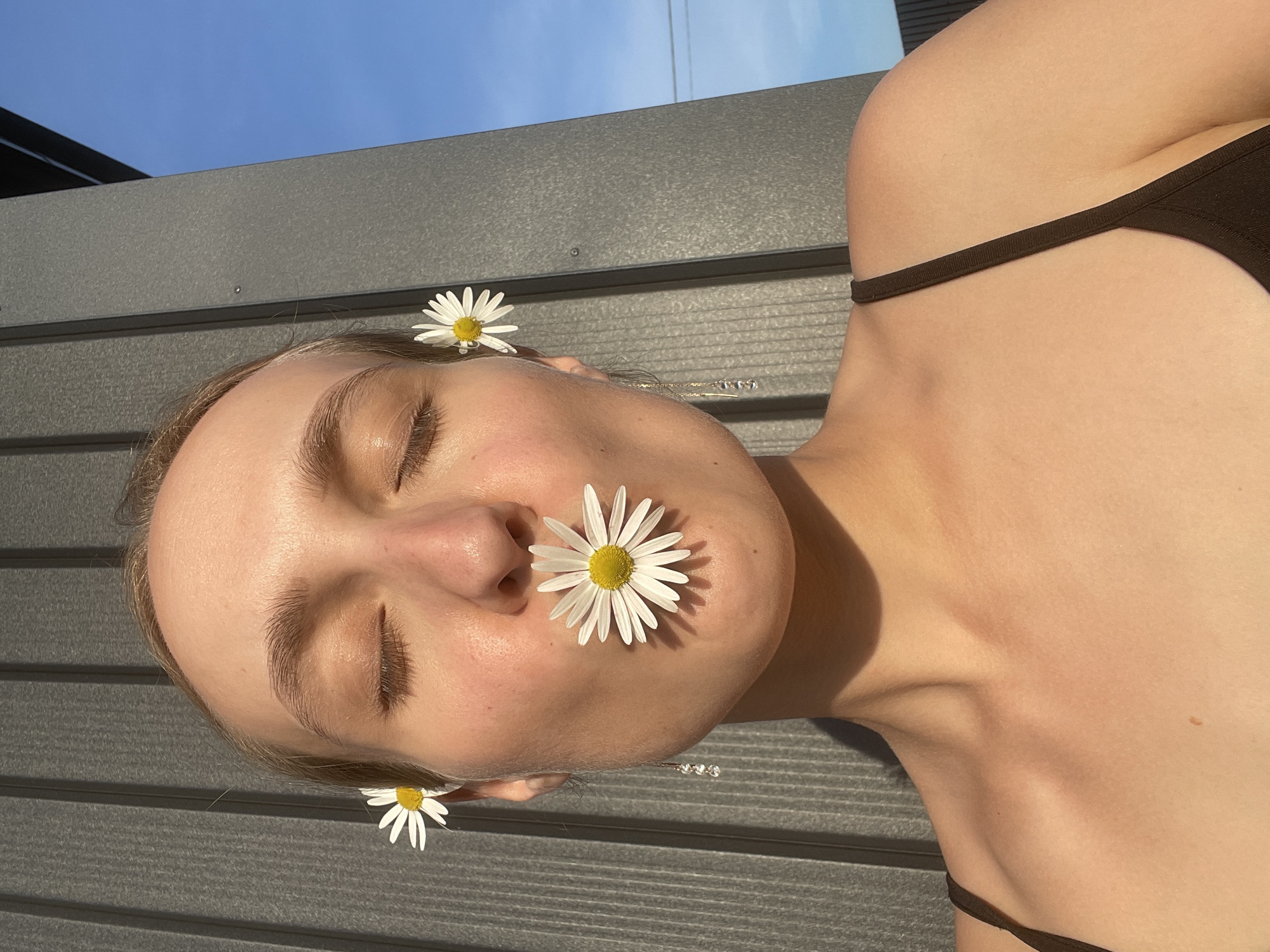 A woman with fair skin is lying on a gray wooden surface outdoors. She has three daisy flowers placed on her face: one on her forehead, one by her ear, and one on her nose. She is wearing a black top with thin straps. The background shows a blue sky and a section of a building or fence. The location is Podshivalovo, Russia.