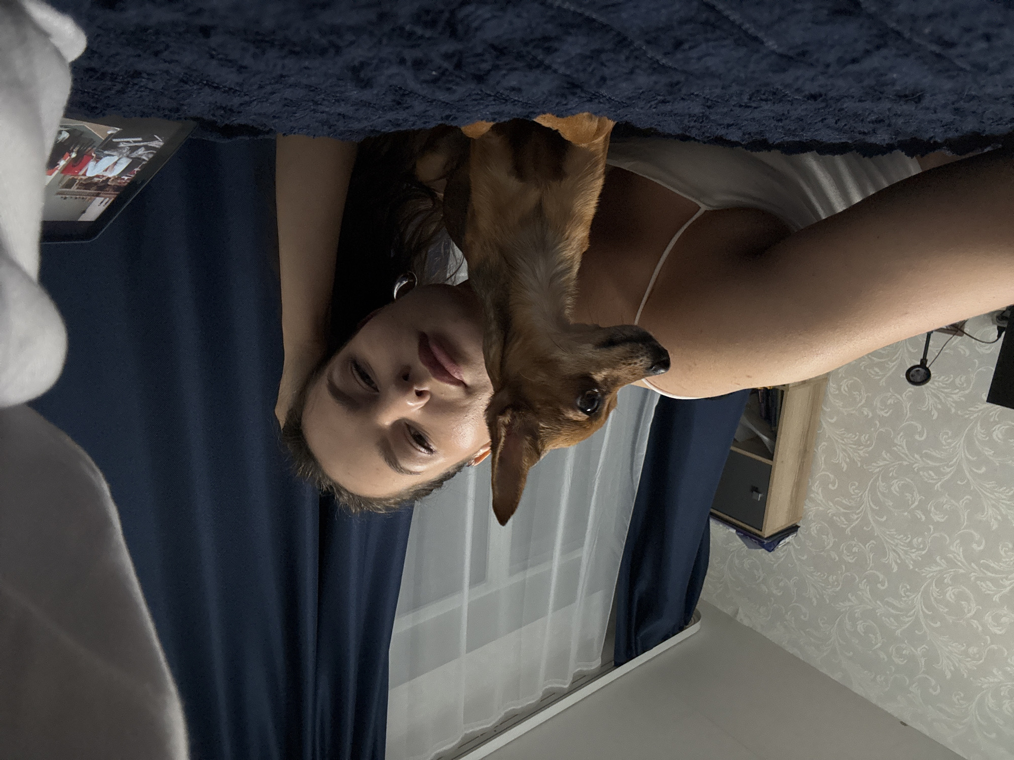 A young woman is lying in bed with a small brown dog. She is on her left side, with her head propped up on her left arm. The dog is in front of her, facing to her left. She is wearing a white tank top. The bed is covered with a dark blue comforter. In the background, there are dark blue curtains and a patterned wallpaper. A tablet is visible on the right side of the bed, displaying a group of people. The scene appears to be indoors, likely a bedroom in an apartment.