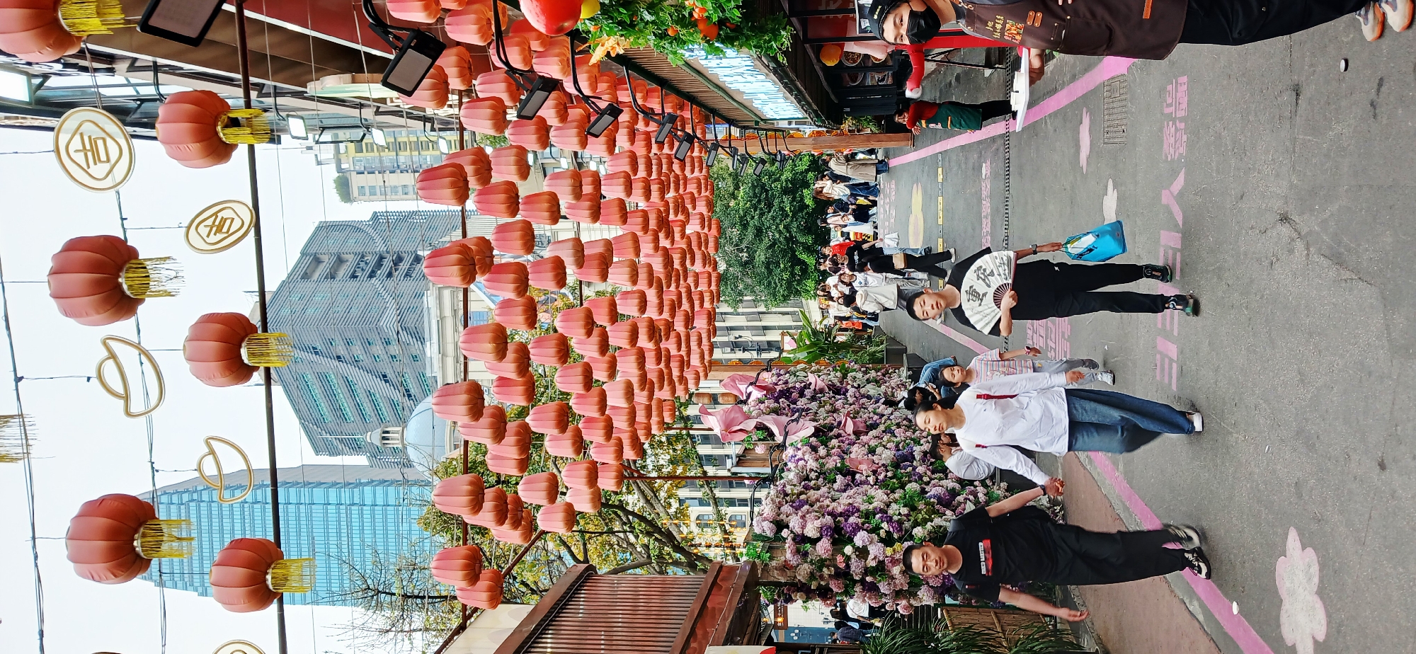 The image captures a bustling street scene in Yuzhong District, China. Rows of pink lanterns hang overhead, stretching down a paved path lined with buildings. A crowd of people stroll along the path, and some are engaged in conversations.
A variety of commercial establishments frame the edges of the path. In the background, a modern glass skyscraper stands in contrast to the traditional decorations.
The foreground includes people in modern casual clothing. One person is holding a traditional fan. The pavement has painted cherry blossoms and other designs. The presence of food vendors suggests a possible market or festival atmosphere.