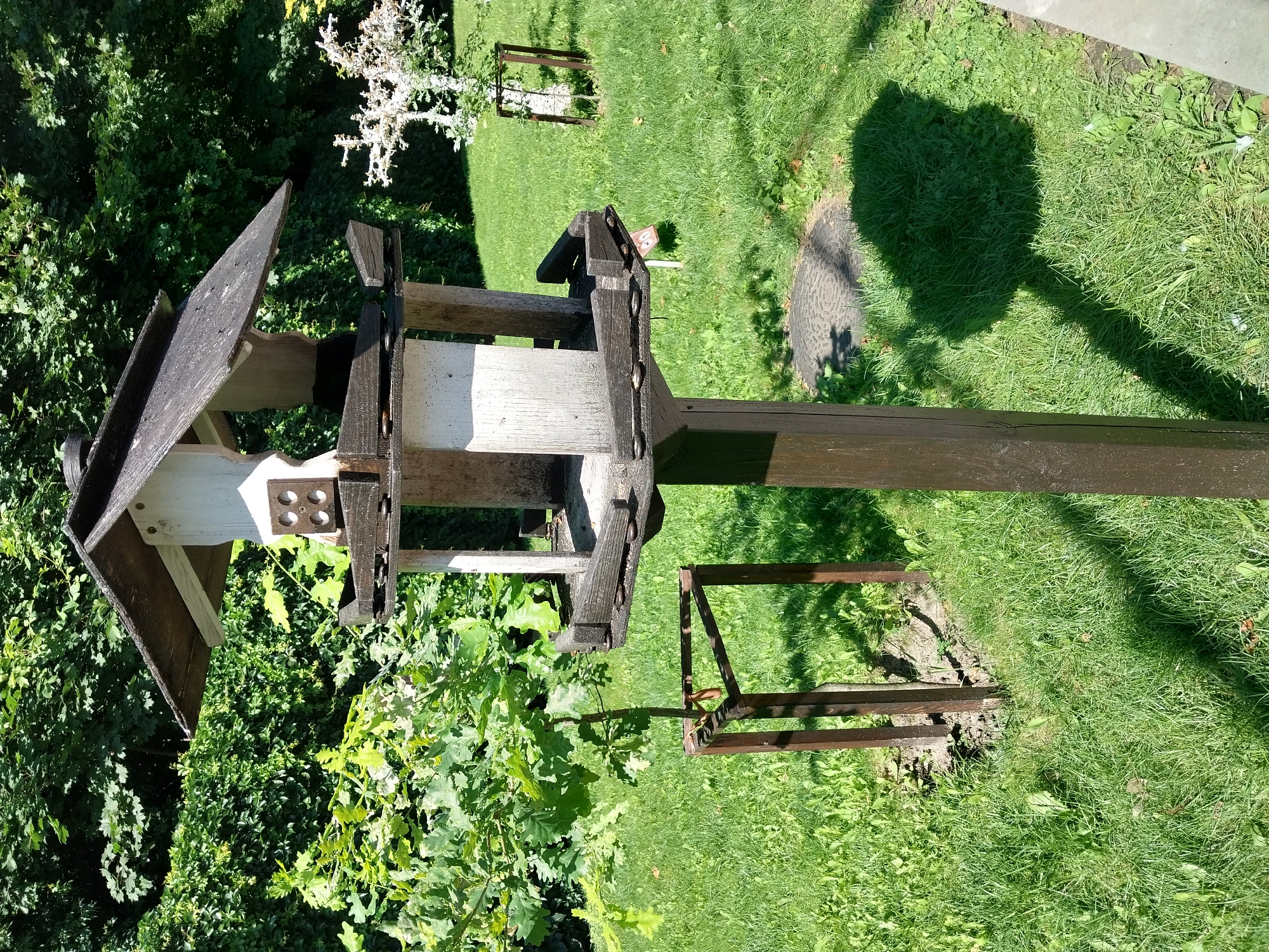 This is a medium shot, taken outdoors on a sunny day, of a wooden birdhouse on a pole in a grassy yard. The birdhouse is multi-leveled and appears weathered, with a dark roof and white body. It has several compartments and feeders. The pole supporting the birdhouse is thick and dark brown. In the foreground, the grass is green and well-maintained, with some scattered leaves. To the left of the birdhouse, a young tree is staked with a brown wooden frame. In the background, lush green foliage and trees form a dense backdrop. A small, bare-branched tree with white accents is visible to the right, also with a stake. A circular manhole cover is partially visible on the ground in front of the birdhouse. The overall scene suggests a park or a backyard in a residential area, likely in the city of Dnipro, Ukraine, given the context of the prompt. The lighting suggests it is daytime, with shadows cast by the birdhouse and surrounding objects.