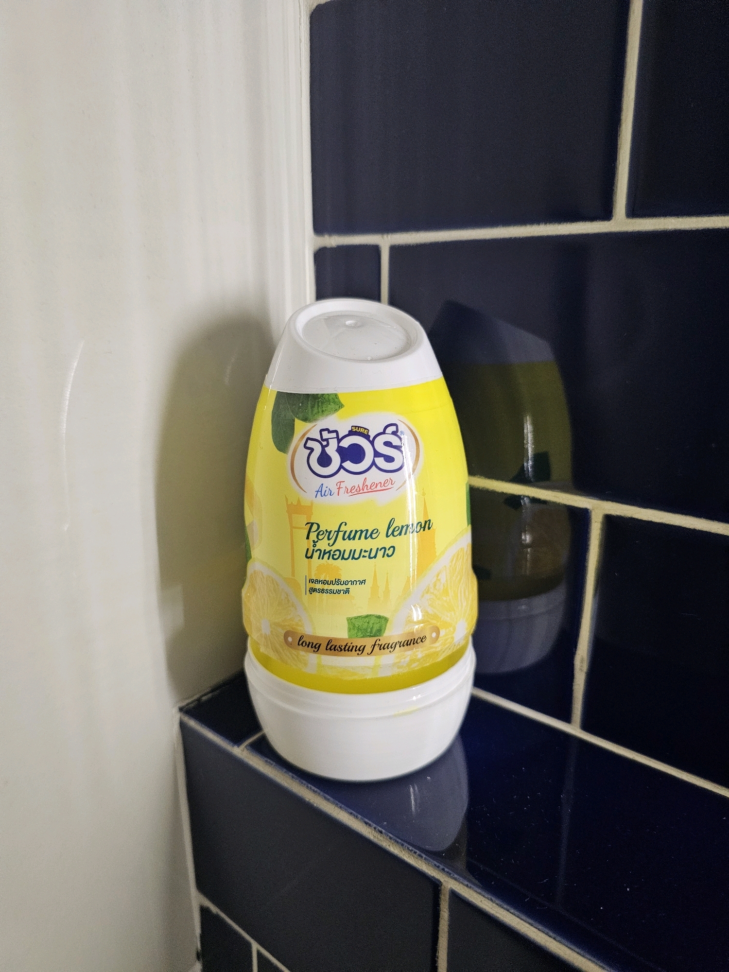 A yellow and white plastic air freshener bottle with a "SURE 30" logo and "Air Freshener" text is positioned on a dark blue tiled shelf in what appears to be a bathroom. The bottle's label features images of lemons and green leaves, with text in English and Thai, including "Perfume lemon" and "long lasting fragrance." The surrounding environment consists of a light-colored wall on the left and dark blue subway tiles with light grout in the background and on the shelf. The reflection of a green object is visible on the dark tiles behind the air freshener. This is a still life composition, likely photographed in a residential bathroom setting in Bangkok, Thailand.