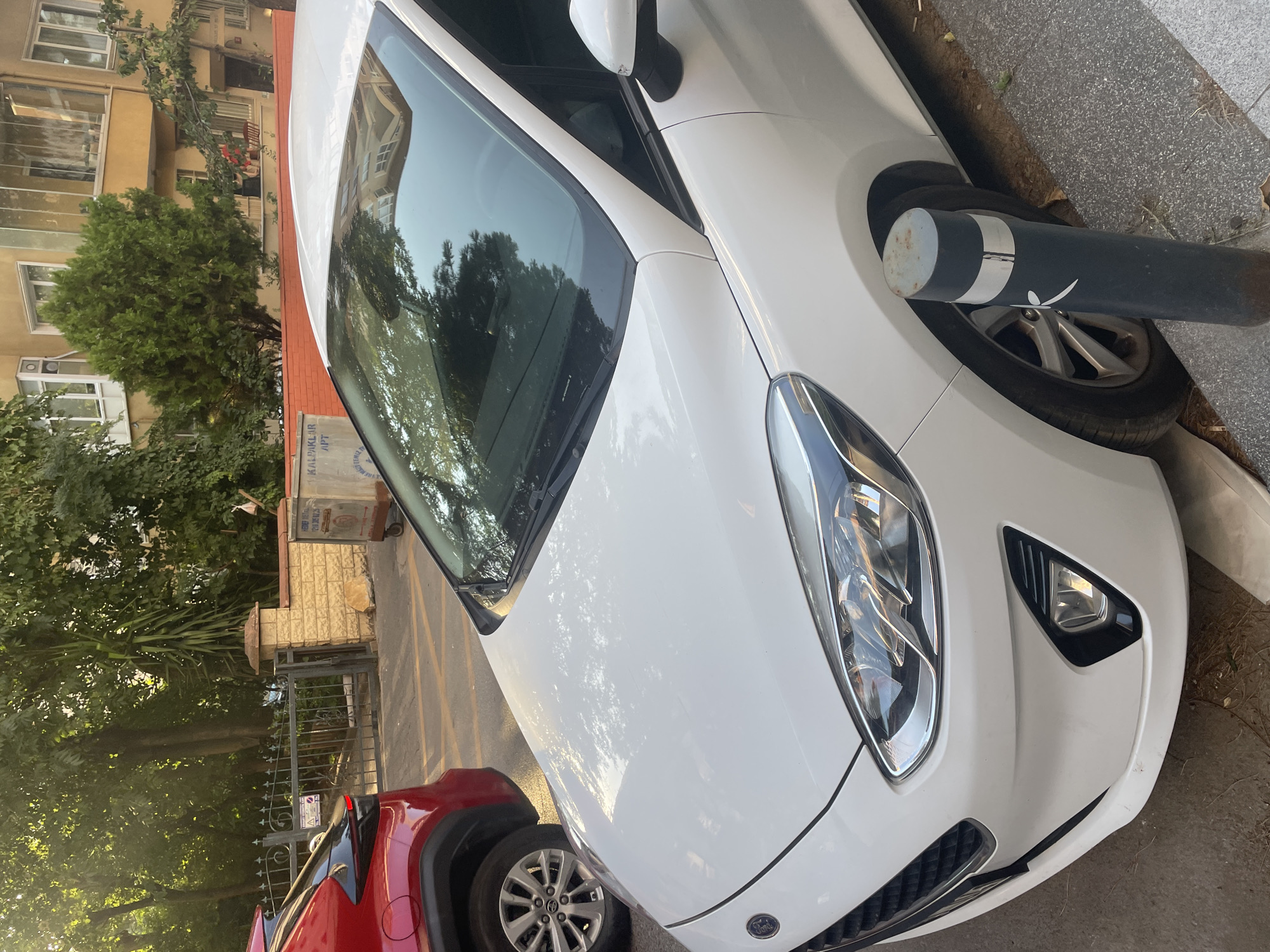 A white Ford Fiesta is parked on a street in Kadıköy, Turkey. The car is facing the viewer and is slightly angled to the right. It is a sunny day, and the car is reflecting the trees and buildings around it. The car is parked next to a red SUV. In the background, there is a building with yellow walls and a glass-enclosed balcony. There is also a metal fence with a gate and a brick wall. A black bollard with white reflective stripes is in front of the car's front passenger wheel. The street appears to be paved with asphalt and concrete. There are no people visible in the image.